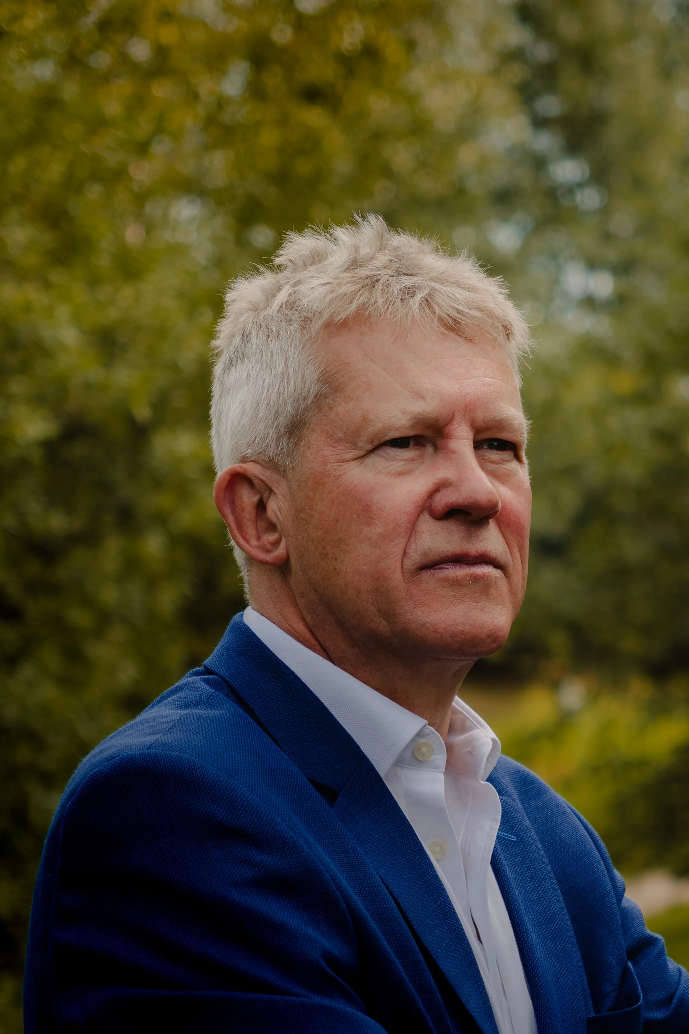 A middle-aged man with gray hair in a blue blazer and white shirt standing outdoors with blurred green trees in the background.
