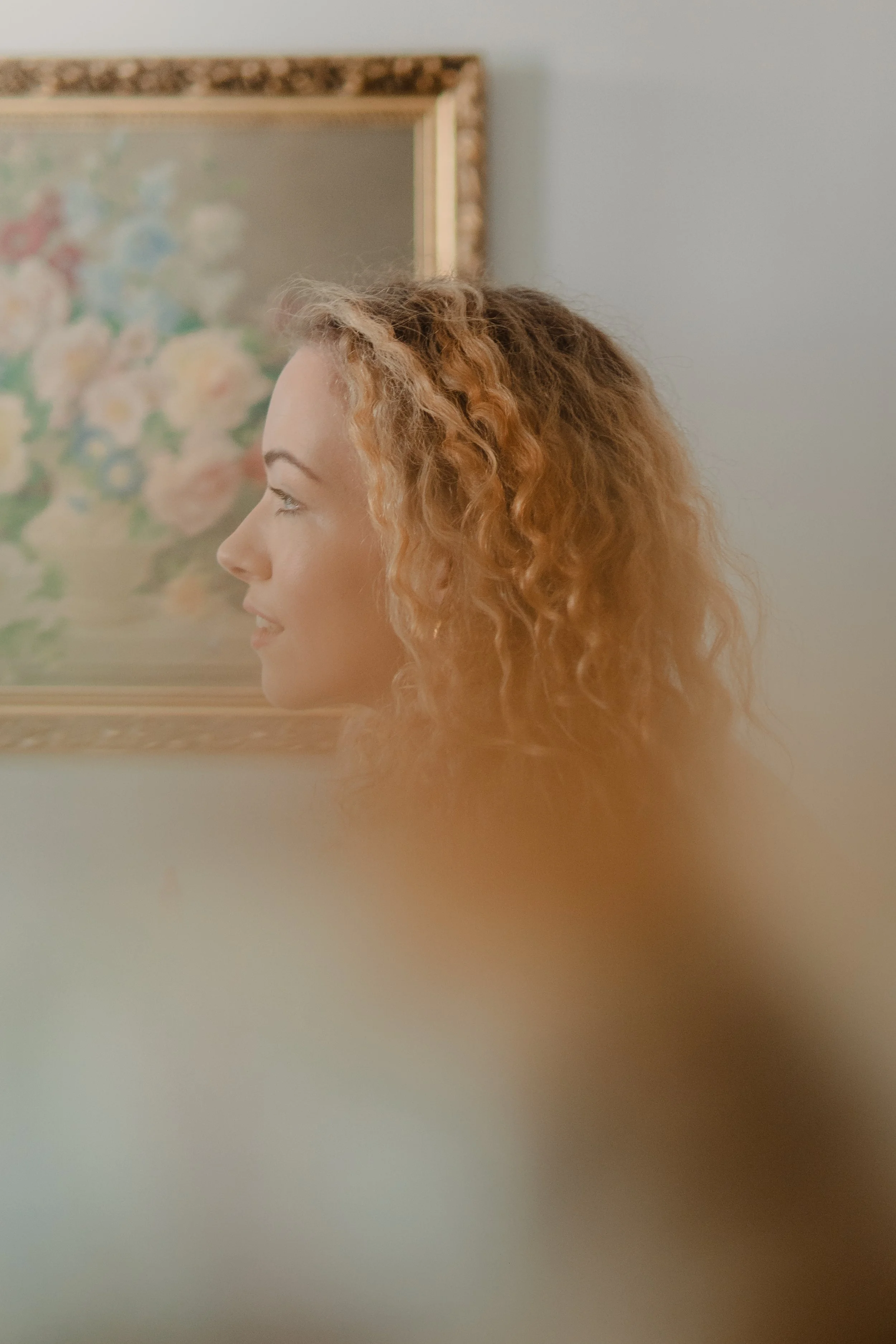 Side profile of a woman with curly, reddish-blonde hair standing next to a floral painting in a softly lit room.