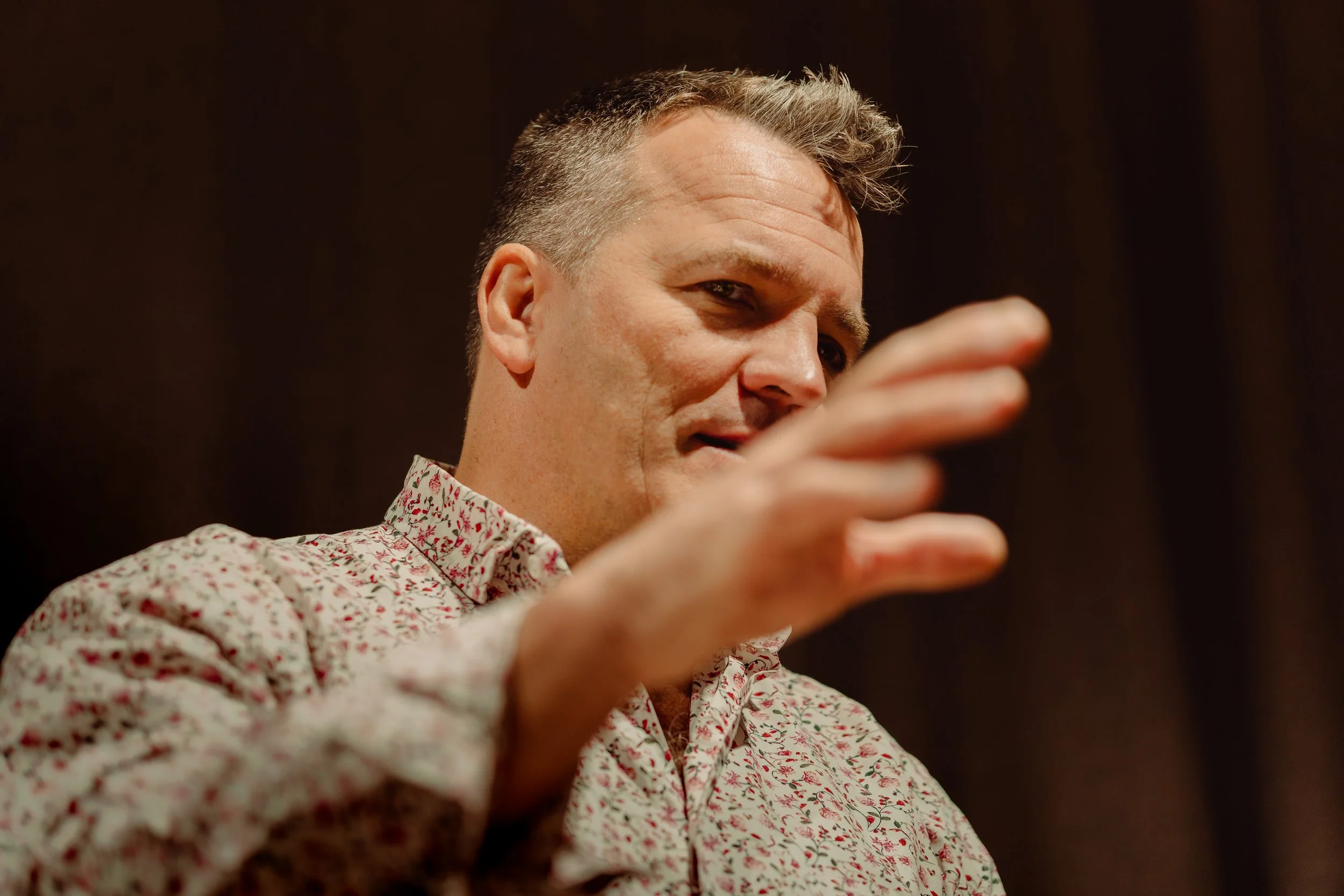 A man with short, styled hair, wearing a light-colored shirt with a floral pattern, reaching out with his right hand while looking slightly downward and to the side against a dark background.