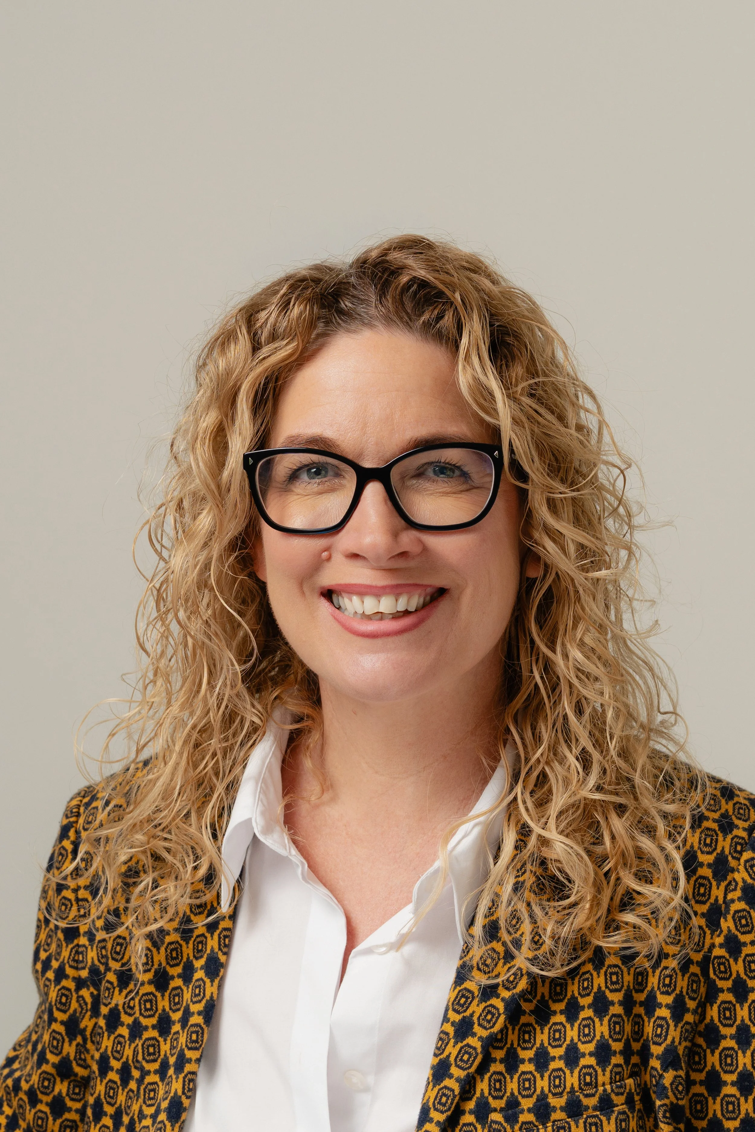 A woman with curly blonde hair wearing black glasses, a white shirt, and a patterned blazer, smiling against a plain background.