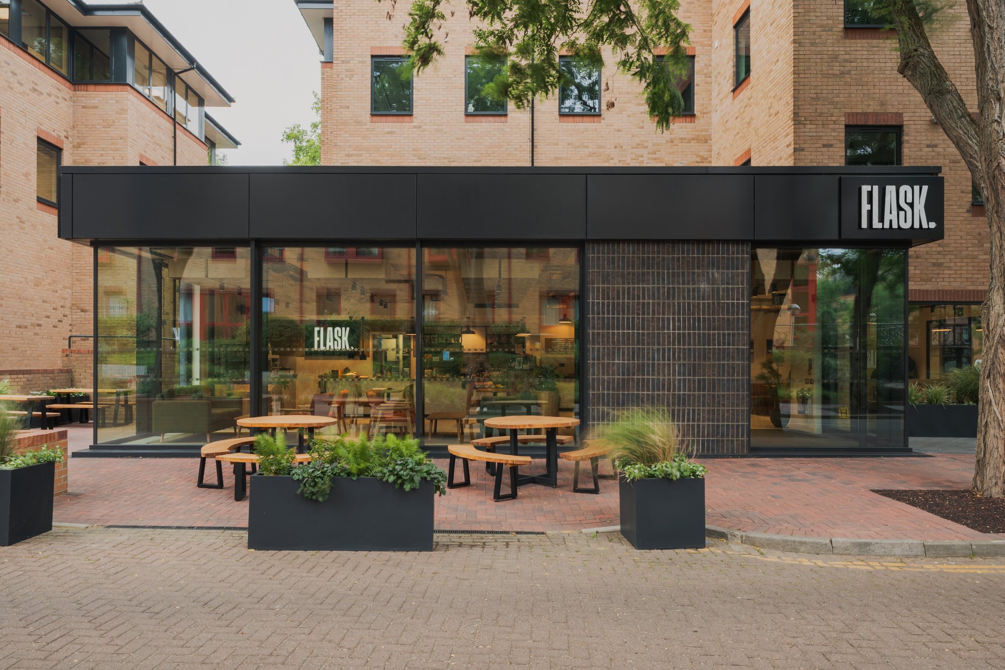 Exterior of a modern building with a small café named FLASK. The café has large glass windows, outdoor seating with round wooden tables and benches, and black planters with greenery. Surrounding the café are tall brick office buildings and trees.