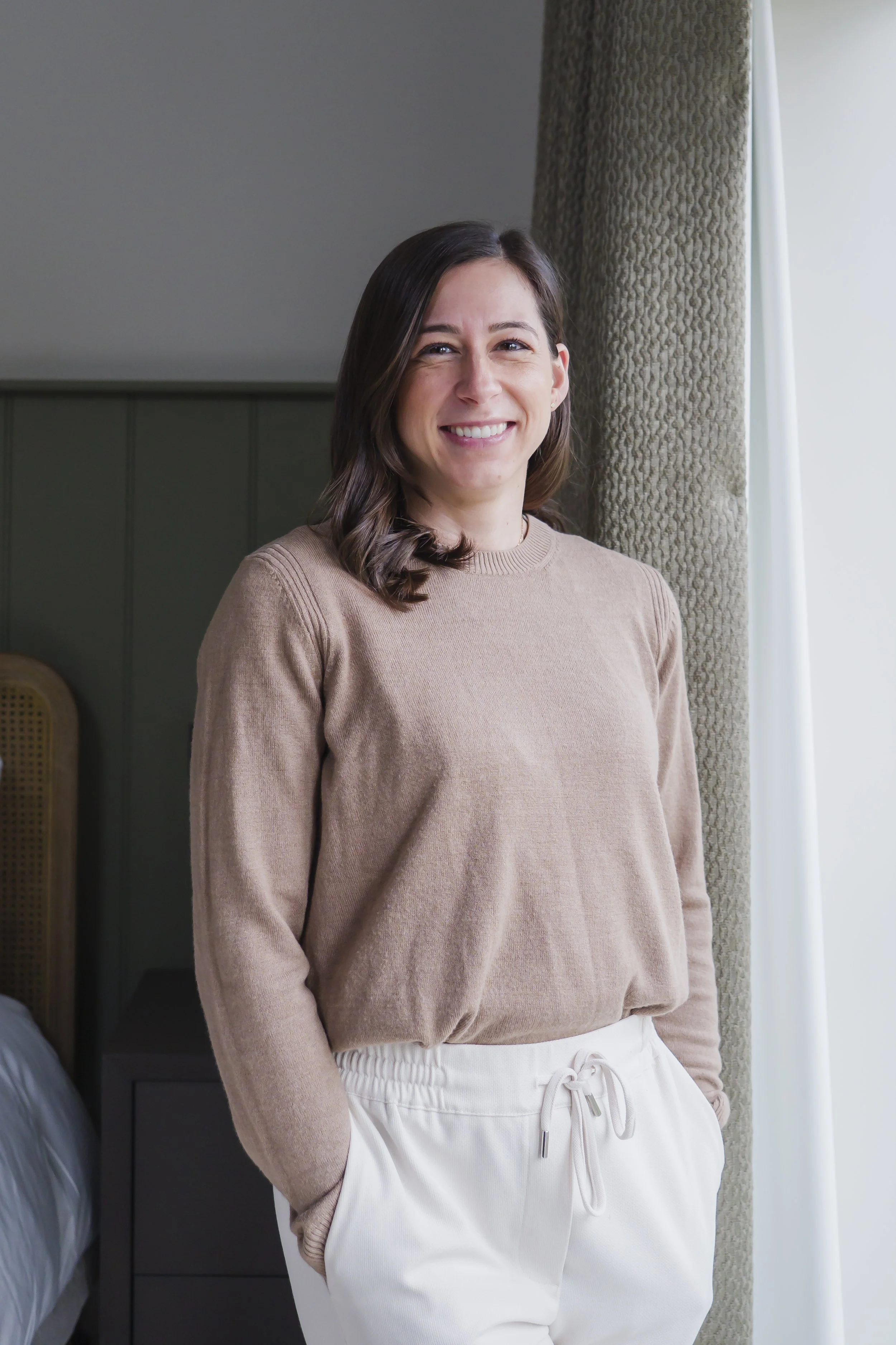 A woman with dark brown hair holding a set of patterned pillows, smiling at the camera, standing in front of a beige wall with decorative wallpaper.