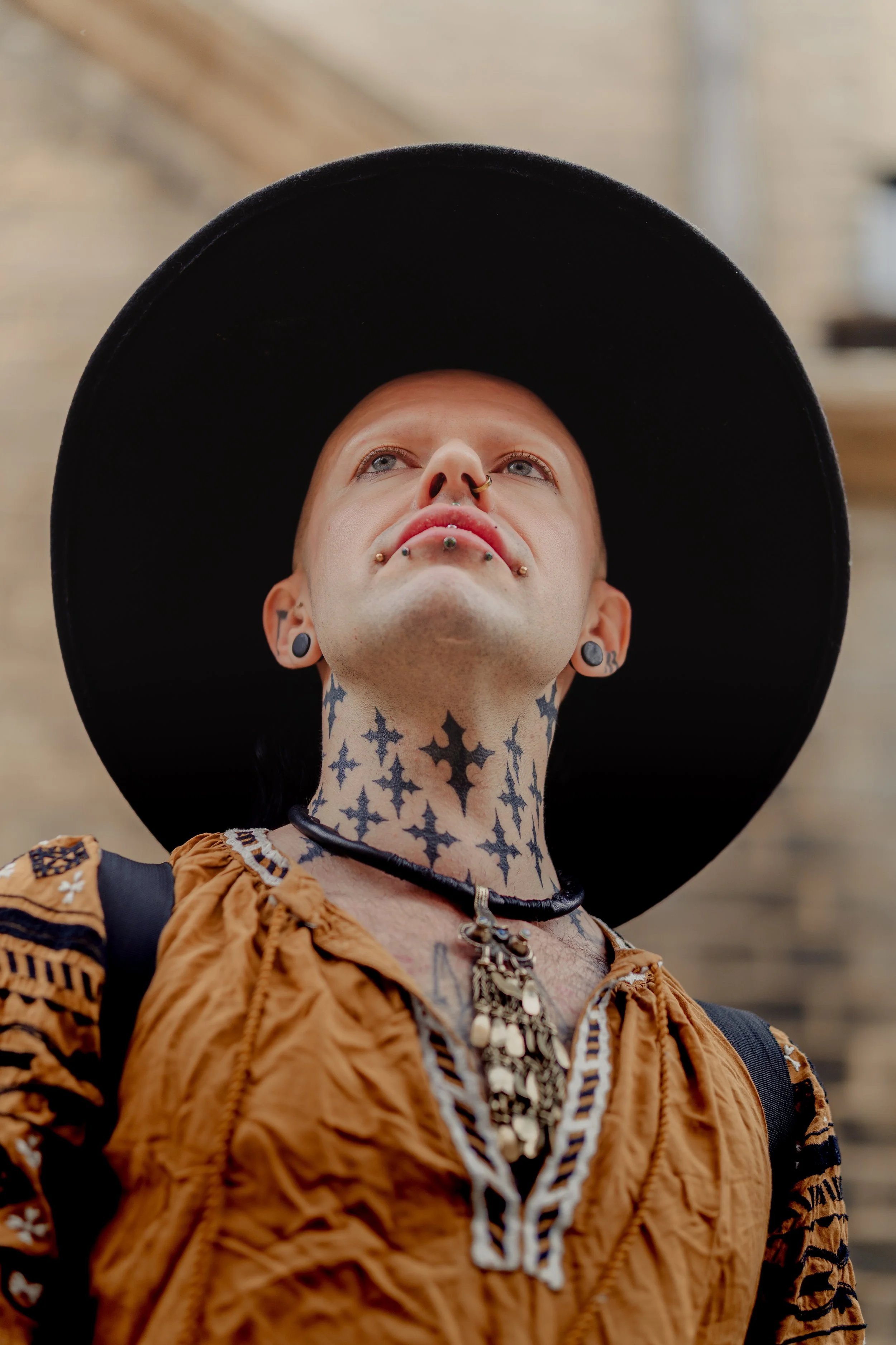 A person wearing a large black hat, with facial piercings and tattoos, looking upward.