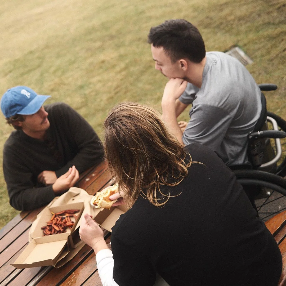 Three people sitting at a wooden picnic table outdoors, with two boxes of food on the table, one containing sweet potato fries; a woman with long hair is eating a sandwich, a young man in a wheelchair is facing her, and another young man wearing a blue cap is talking to them.
