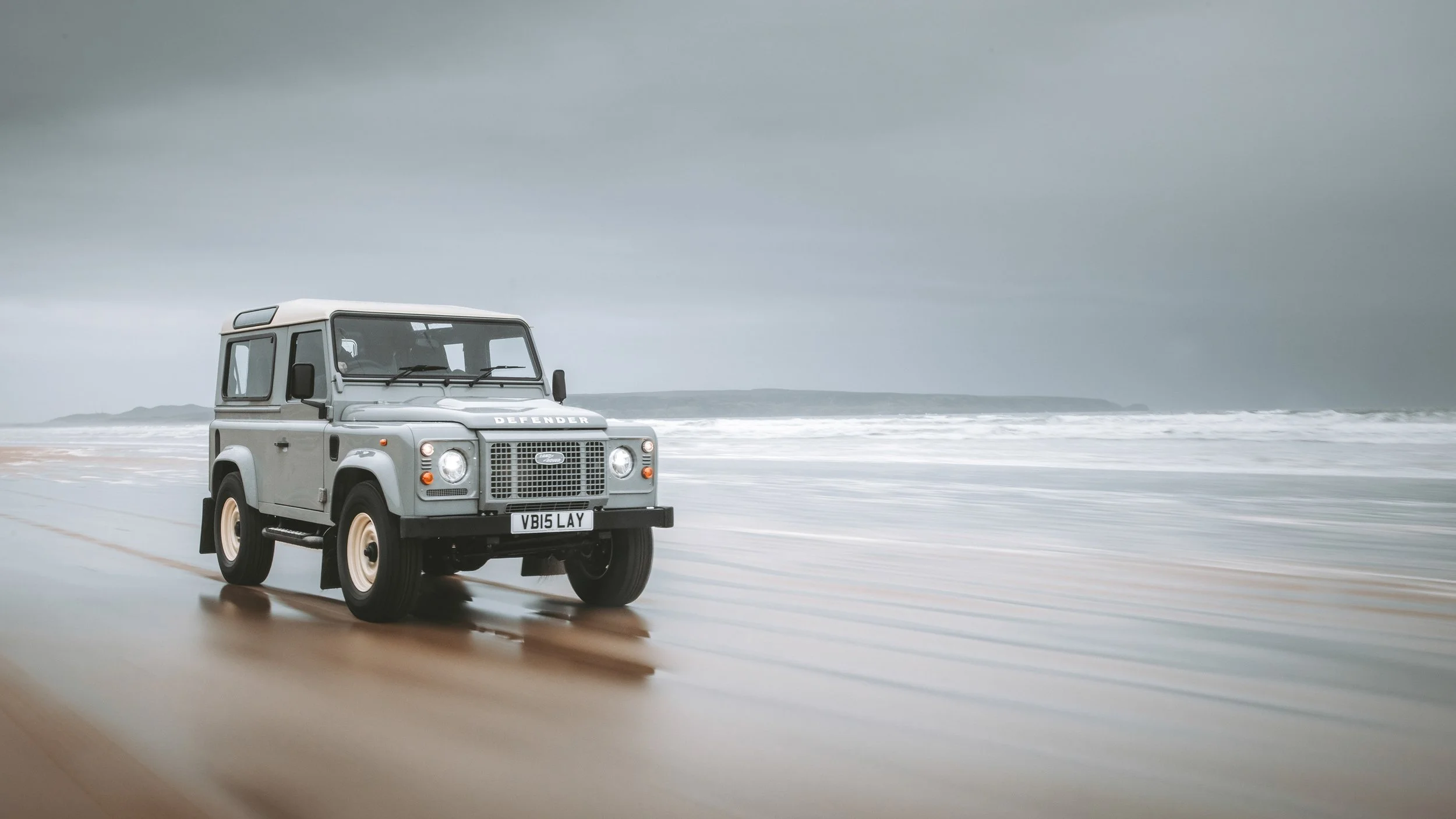 A vintage gray Land Rover Defender driving on a sandy beach with a stormy sky and ocean waves in the background.