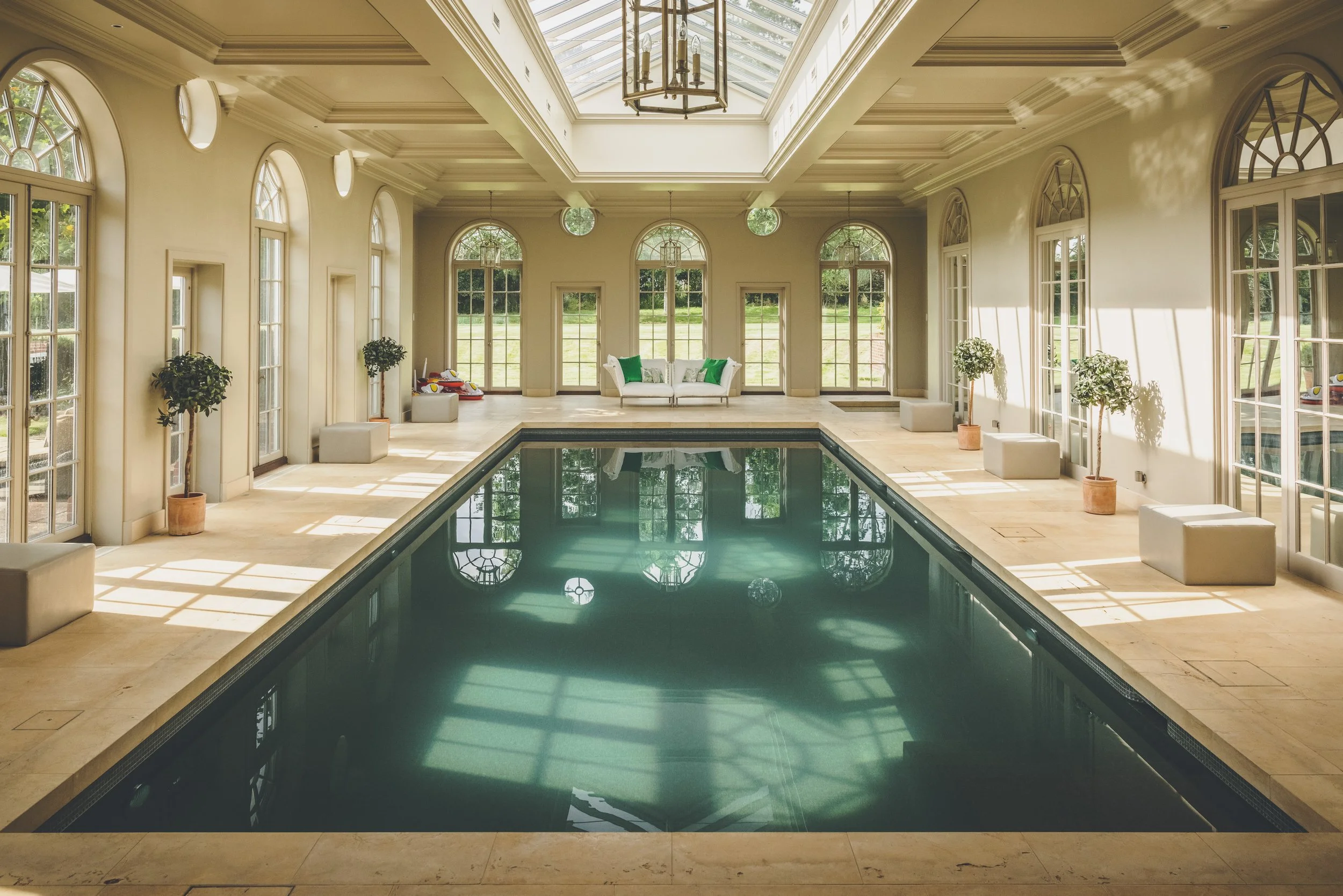 Elegant indoor swimming pool in a bright room with large windows, potted plants, and comfortable seating.