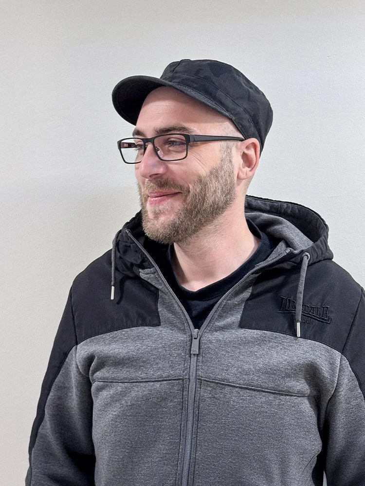 Ein Mann mit Brille, trägt eine schwarze Mütze und eine grau-schwarze Jacke, steht vor einer weißen Wand.