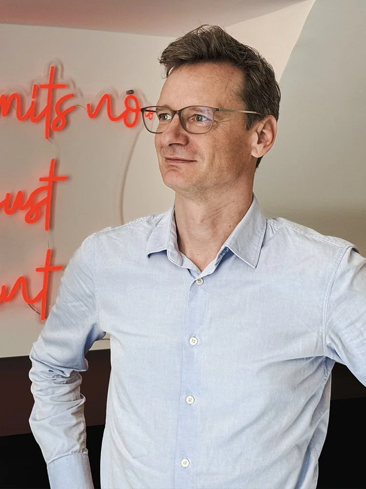 Ein Mann mit Brille in einem hellblauen Hemd steht vor einer weißen Wand mit rotem Neon-Lichtertext.