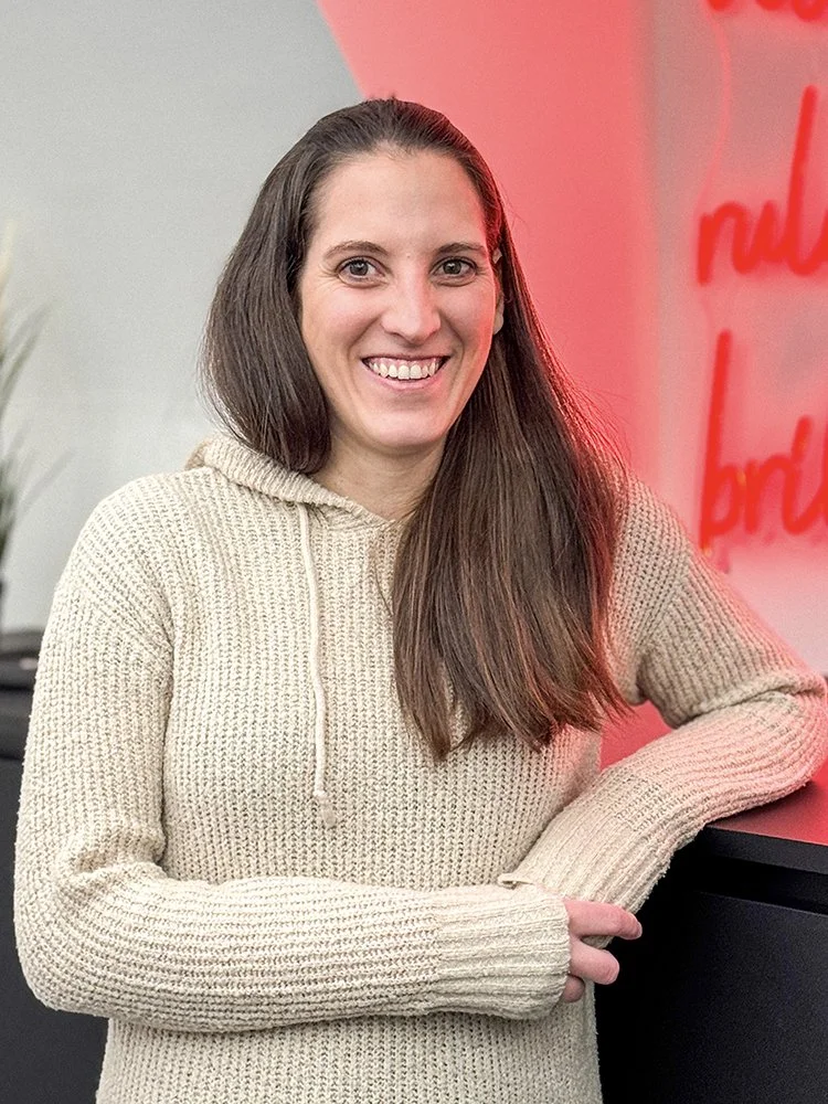 Eine lächelnde Frau mit langen braunen Haaren trägt einen beige Pullover mit Kapuze und steht vor einer roten Neon-Schrift.