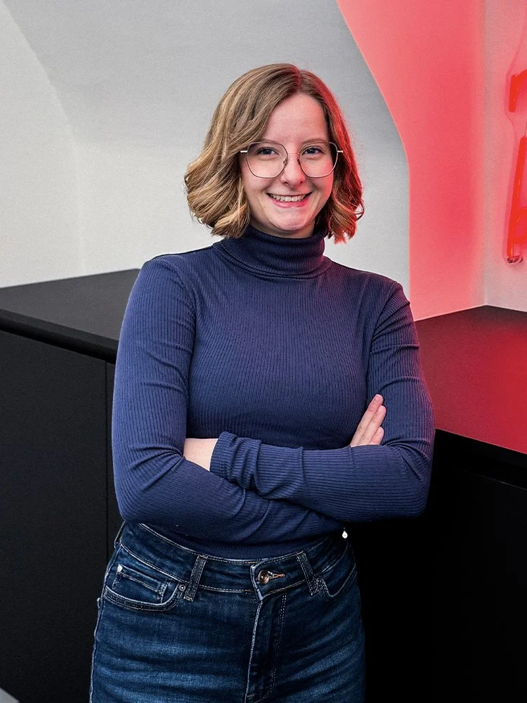 Junge Frau mit Brille, braune Haare, blaue Ripp-Turtleneck, lächelnd mit verschränkten Armen vor schwarzem Möbelstück und roter Neonbeleuchtung im Hintergrund.