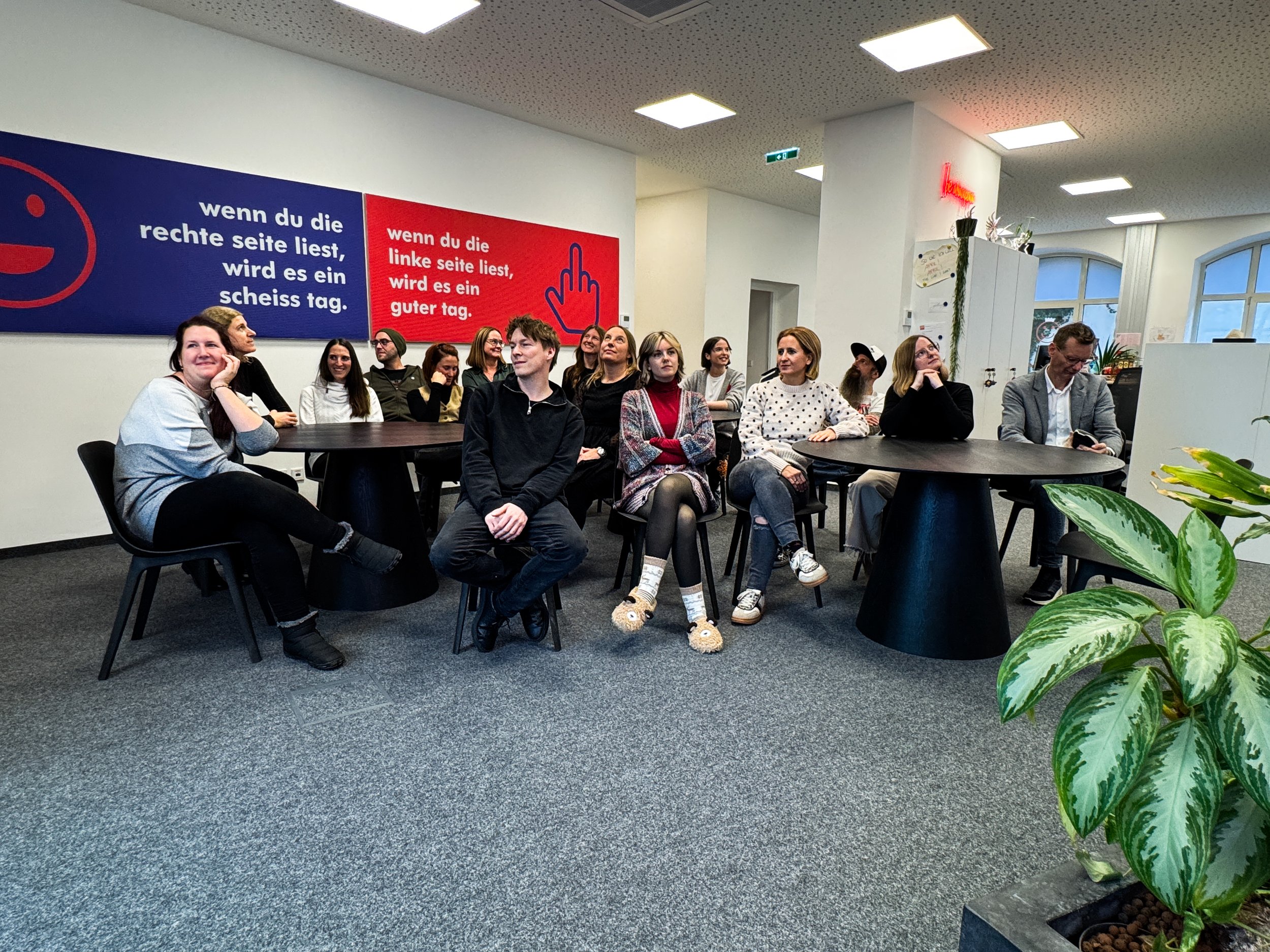 Ein Gruppenfoto von mehreren Menschen, die in einem modernen Büroraum sitzen. An der Wand hängen zwei große blaue und rote Schilder mit deutschen Texten.