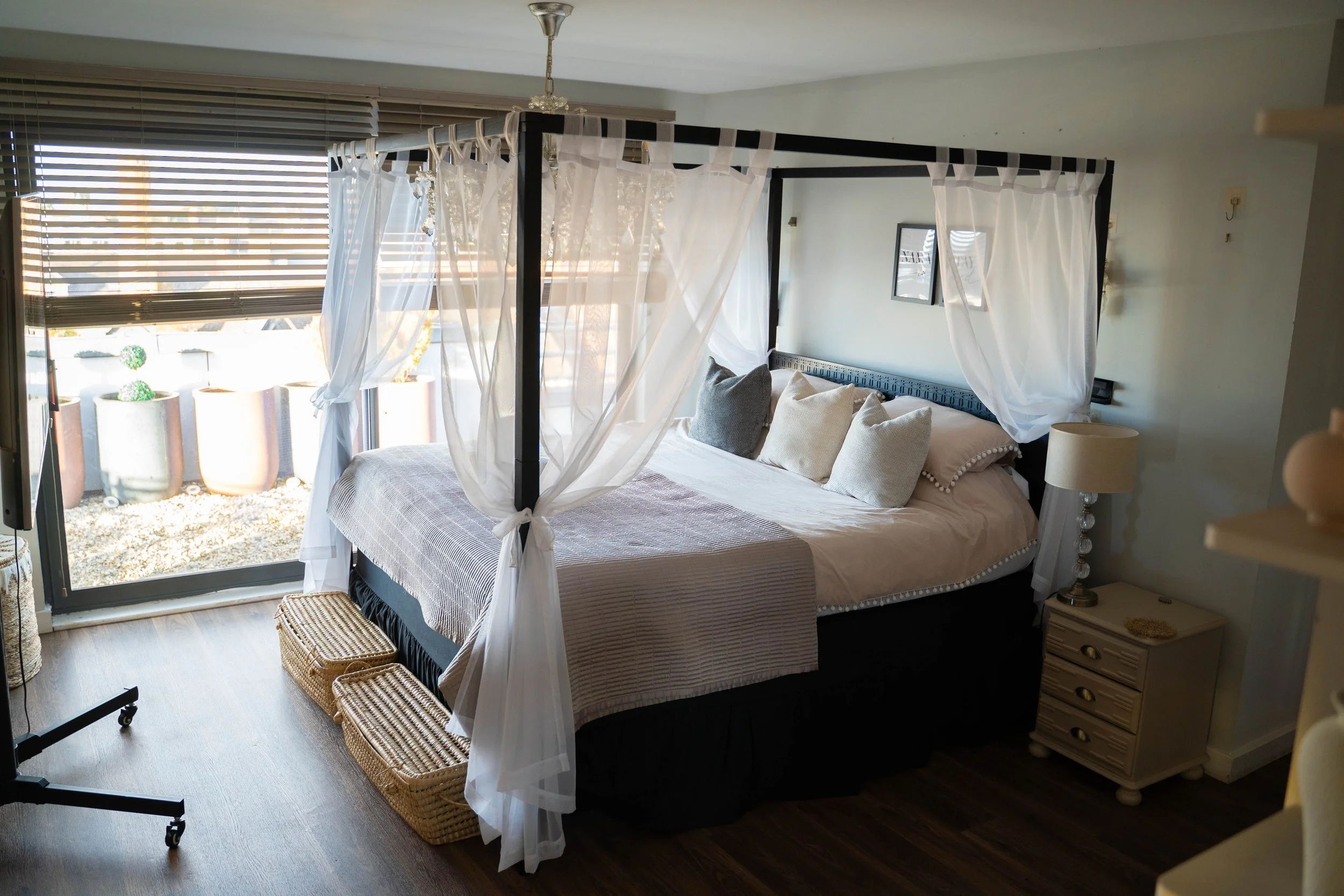 A bedroom with a four-poster bed, white curtains, and a large window with blinds. There are decorative pillows on the bed, a bedside table with a lamp, and a wicker basket on the floor. Outside the window, potted plants are visible.