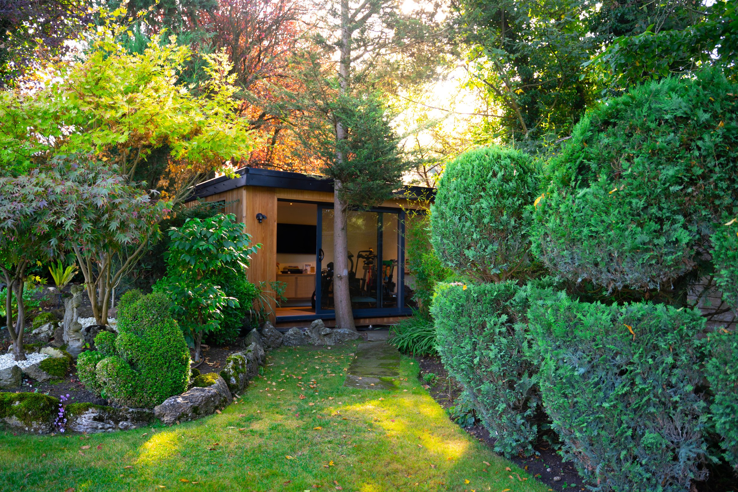 A small backyard garden with neatly trimmed bushes and trees, leading to a modern wooden and glass backyard gym or studio with exercise equipment inside, surrounded by lush greenery and evening sunlight.