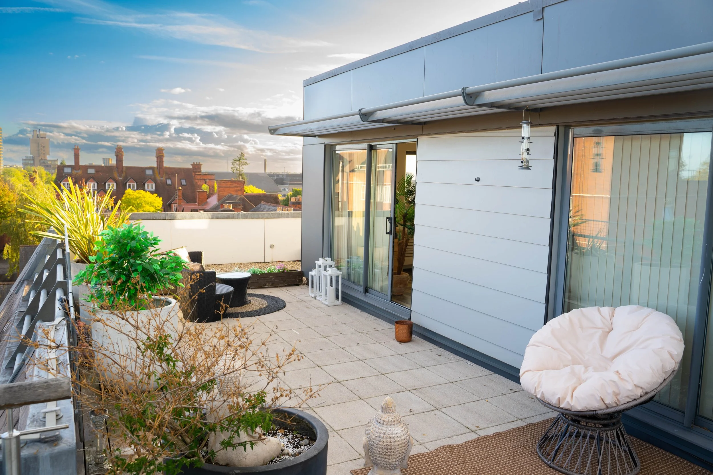 Balcony with potted plants, outdoor seating, lanterns, and a view of red-brick rooftops and a cloudy sky in the distance.