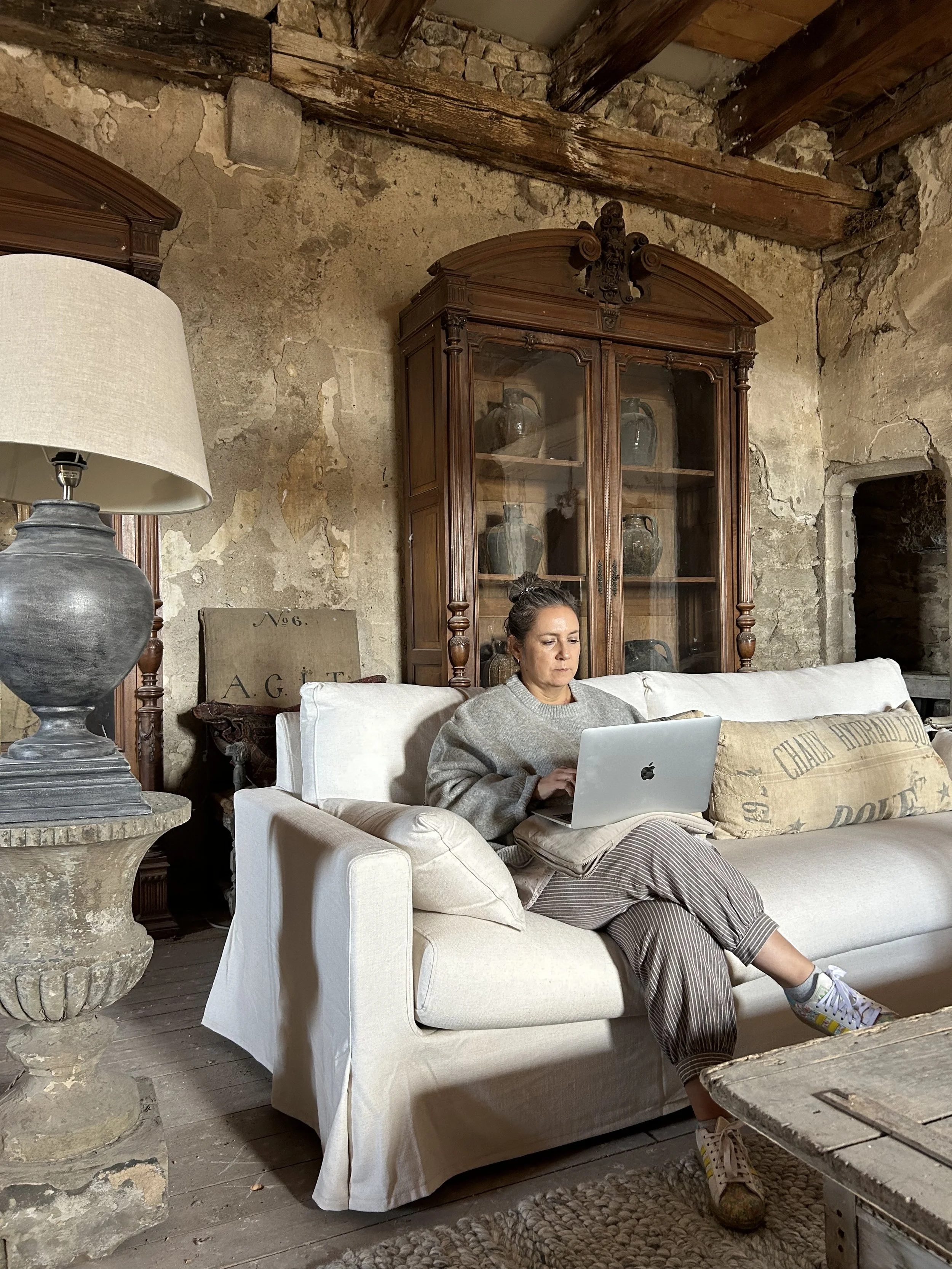 Woman sitting on a cream-colored sofa in a rustic room using a laptop, with a stone wall and wooden ceiling beams in the background.