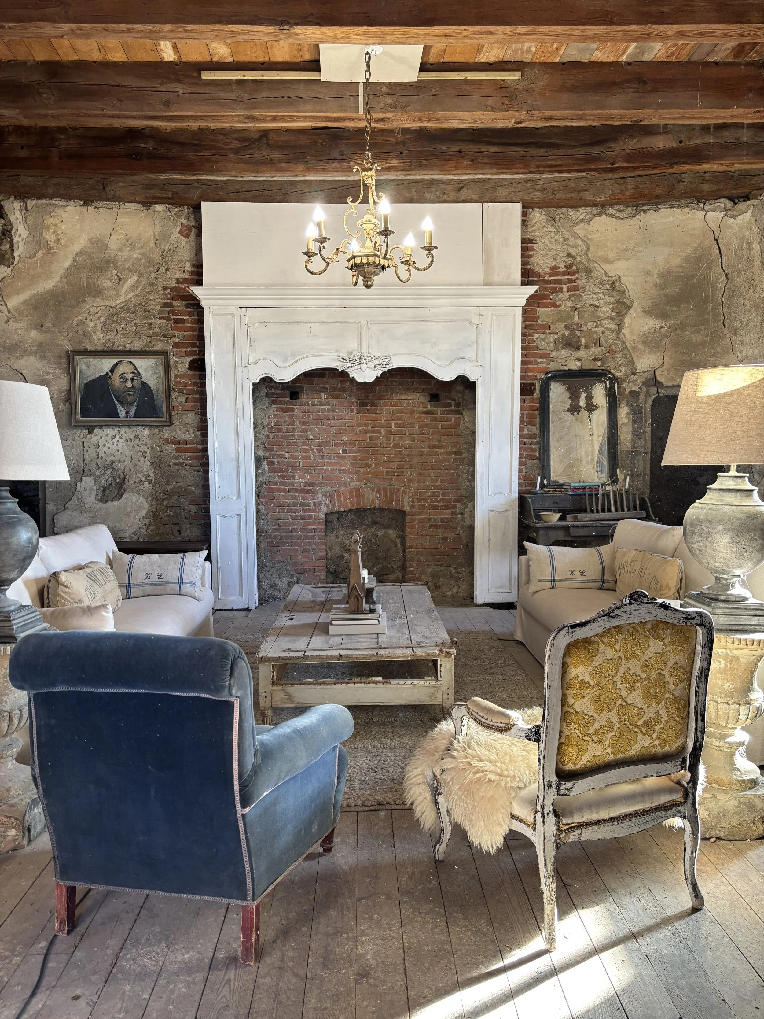 Living room with rustic and vintage decor, featuring exposed wooden ceiling beams, a large white ornate fireplace, a chandelier, two sofas, an armchair with a fur throw, and a distressed coffee table. Walls are a mix of exposed brick and plaster, with a portrait on the left wall and a mirror and painting in the background.