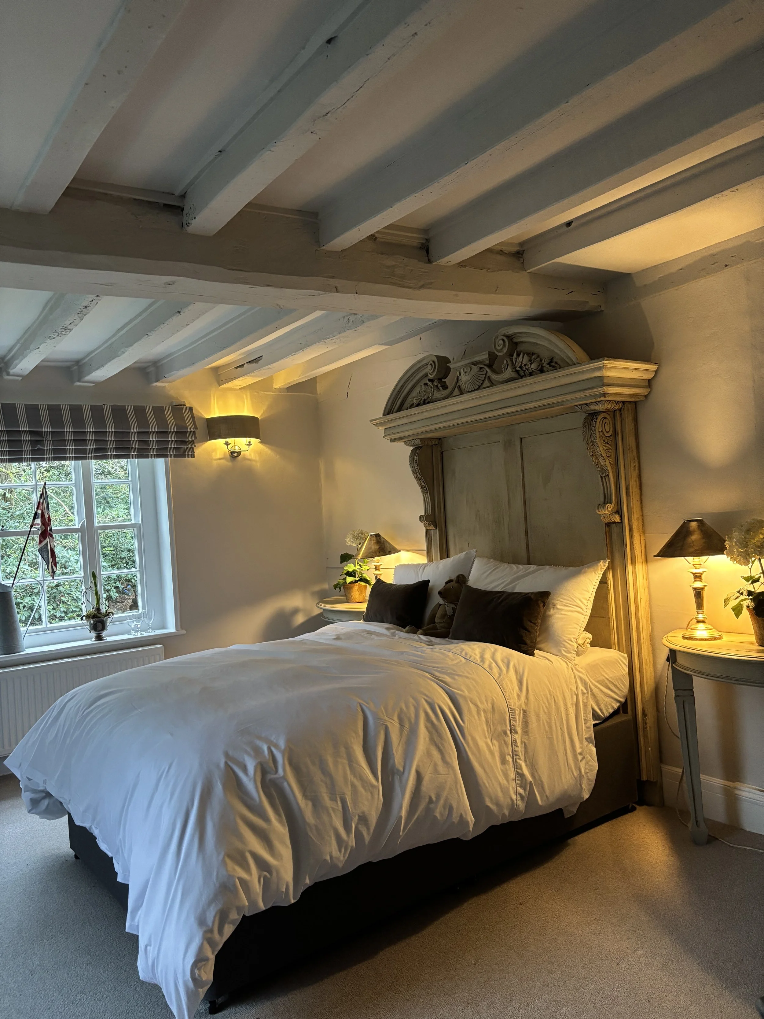 A cozy bedroom with a large bed, white bedding, black and white pillows, bedside lamps, a window with a plaid curtain, and decorative plants, featuring wooden beams on the ceiling.
