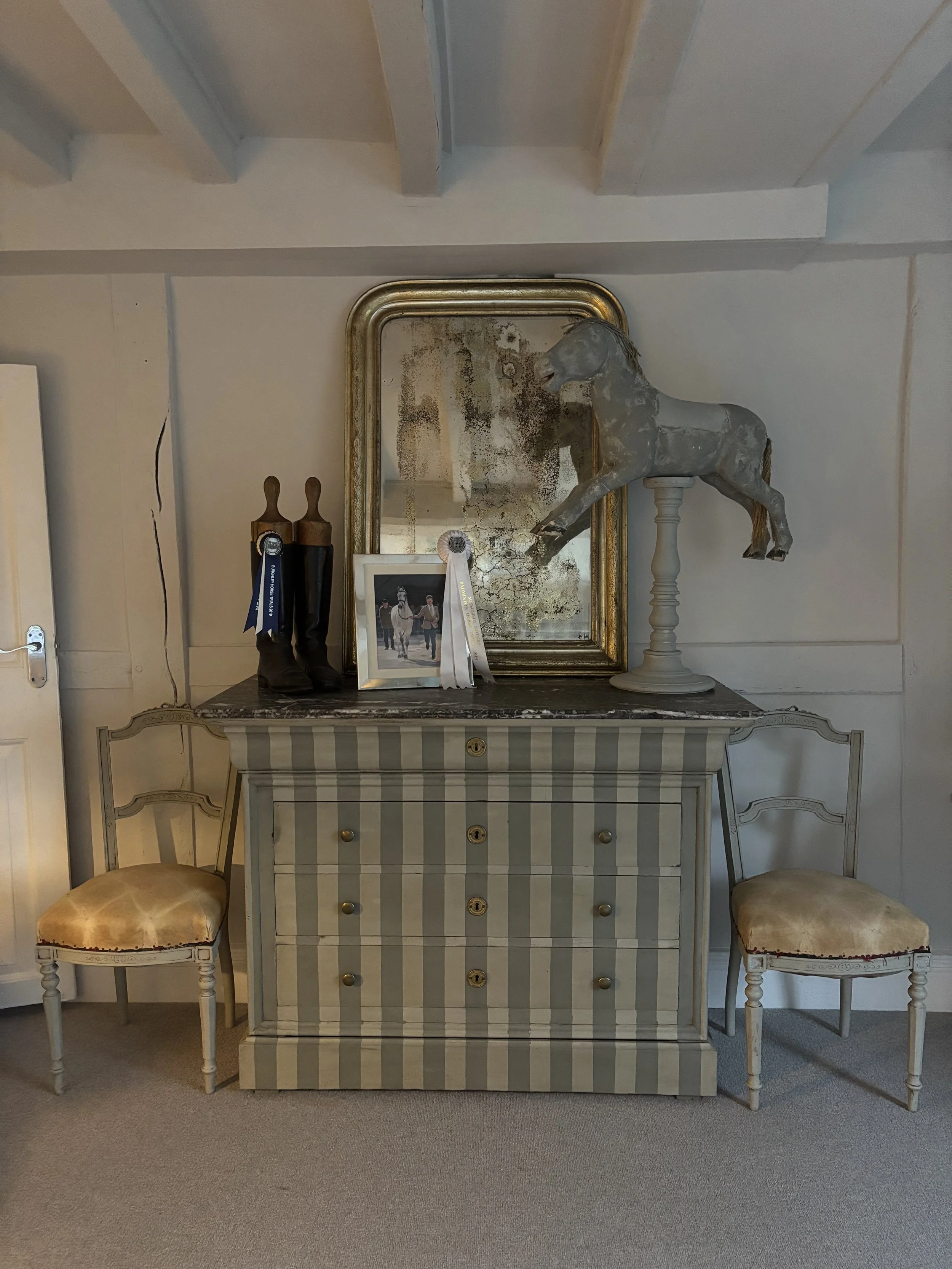 A vintage sideboard with a striped pattern, two chairs on each side, a mirror leaned against the wall, a decorative horse head on a pedestal, a photo frame, and two riding boots with ribbons.