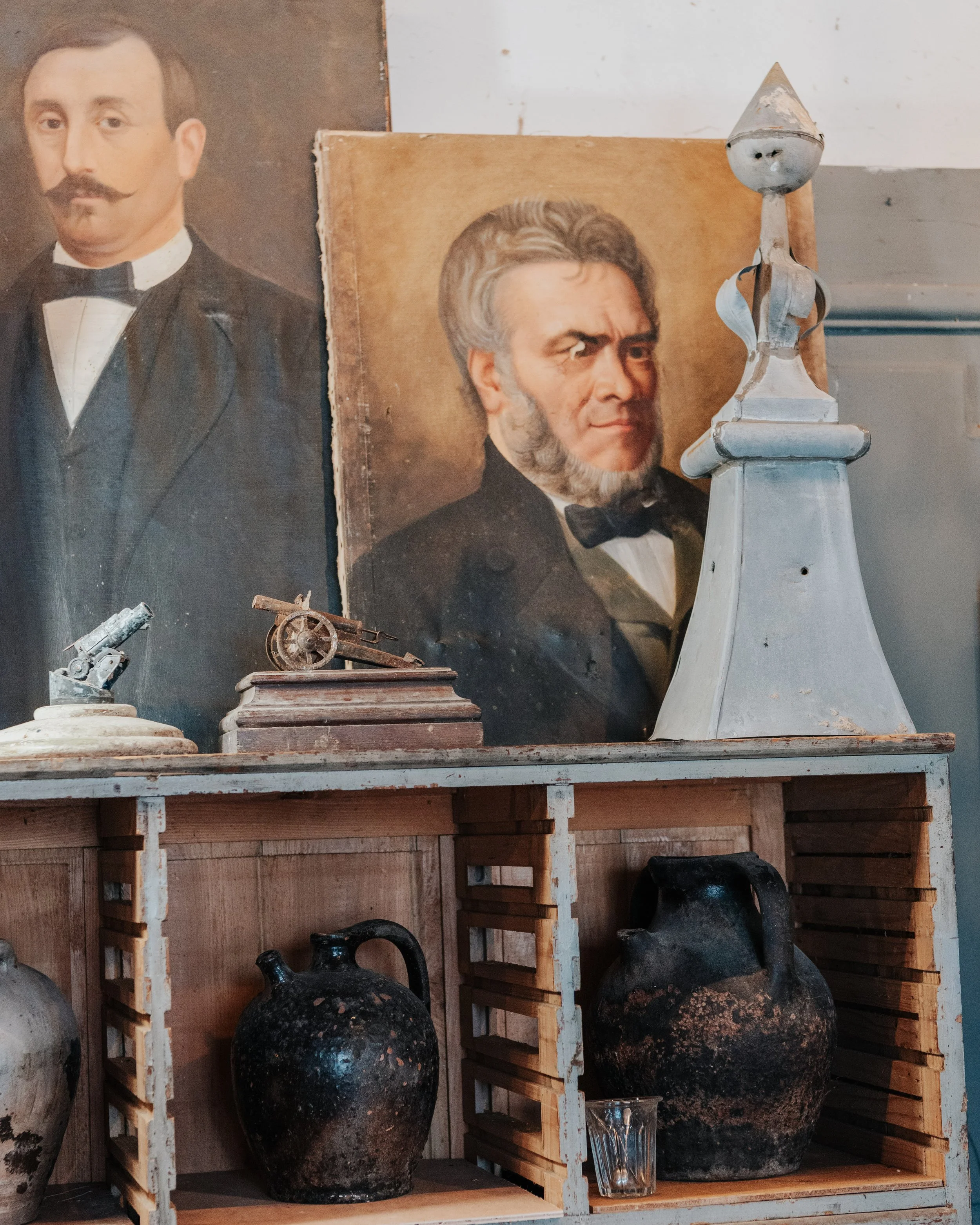 Vintage portraits of two men in formal attire, old pottery jugs, and antique decorative items on wooden shelves.