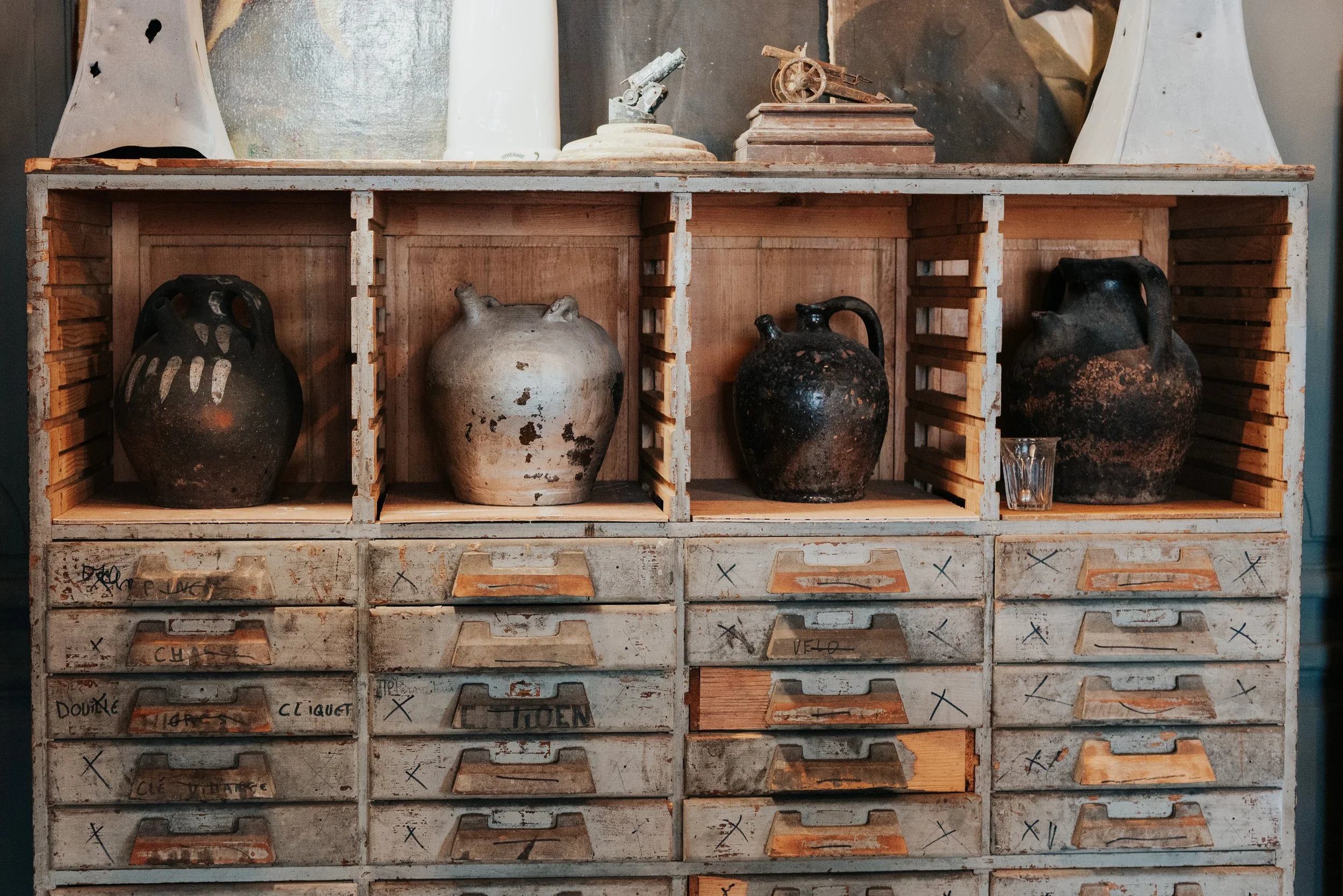 An old wooden storage cabinet with multiple labeled drawers, holding four ancient pottery jugs on top, accompanied by some small decorative items and a glass.