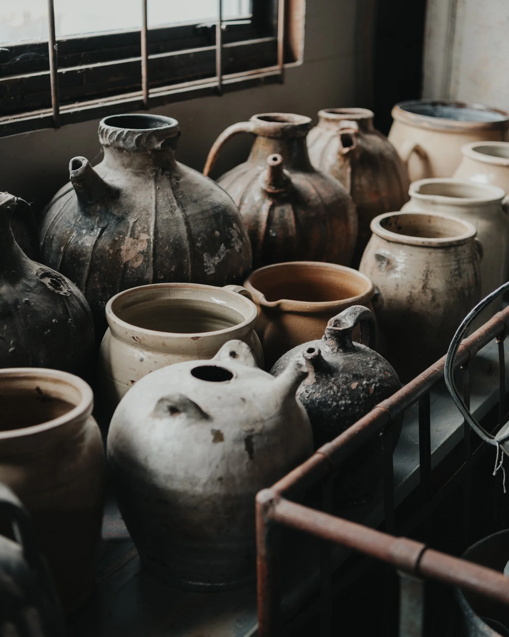 Various old ceramic jugs and pots with weathered textures arranged on a shelf near a window.