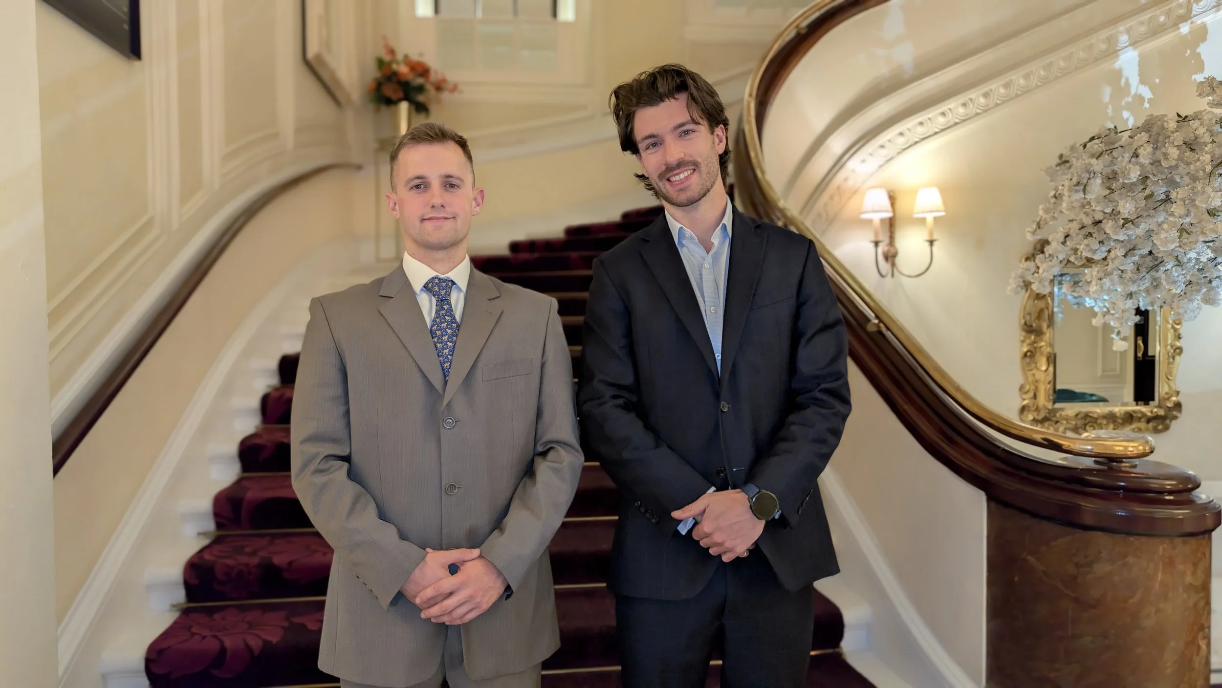 Founders in suits standing on a staircase in an elegant, well-lit interior with decorative wall lighting, a large mirror, and a floral arrangement.