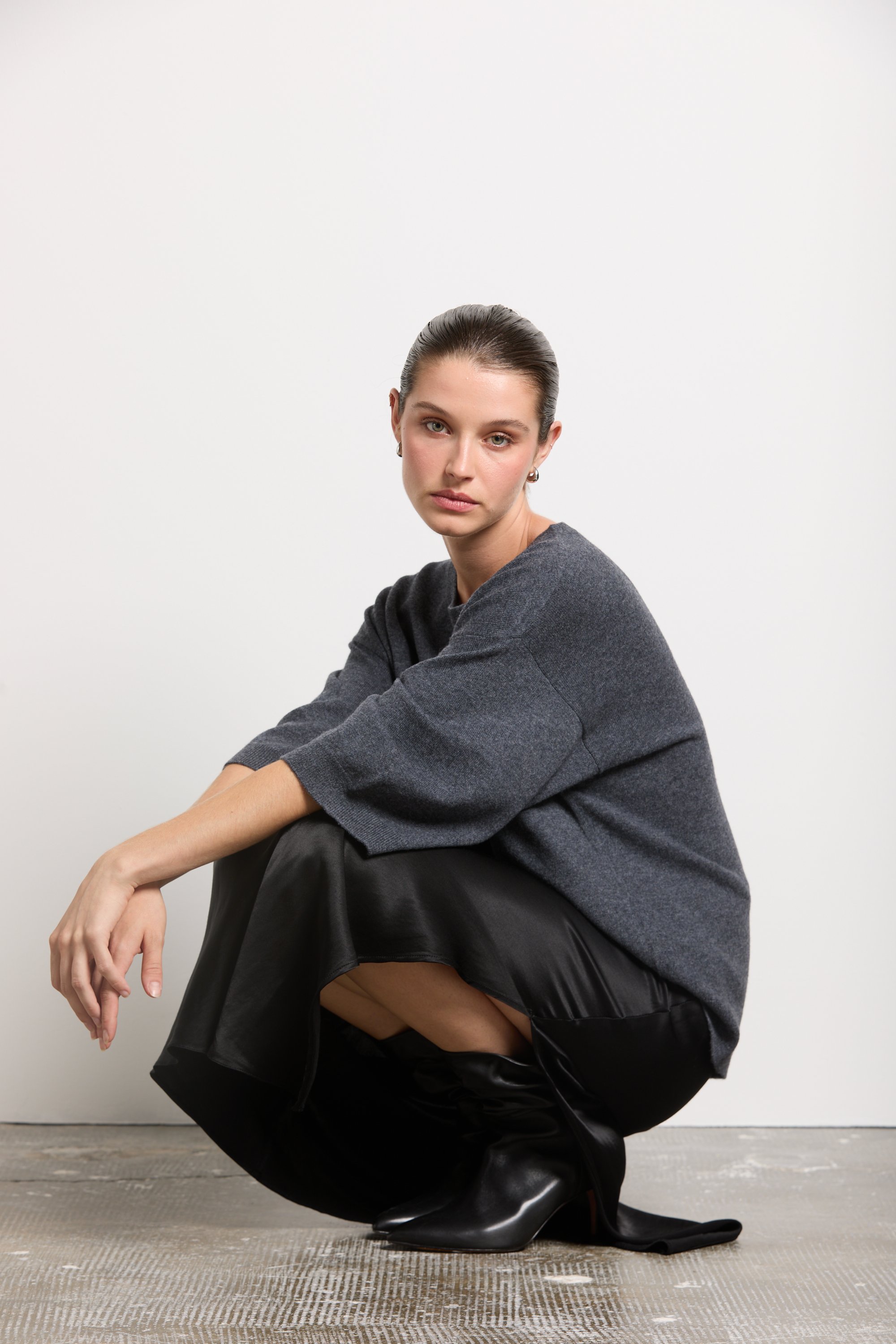 Une femme assise en position accroupie, portant une robe noire en cuir, un pull gris et des bottes noires, avec un fond blanc.