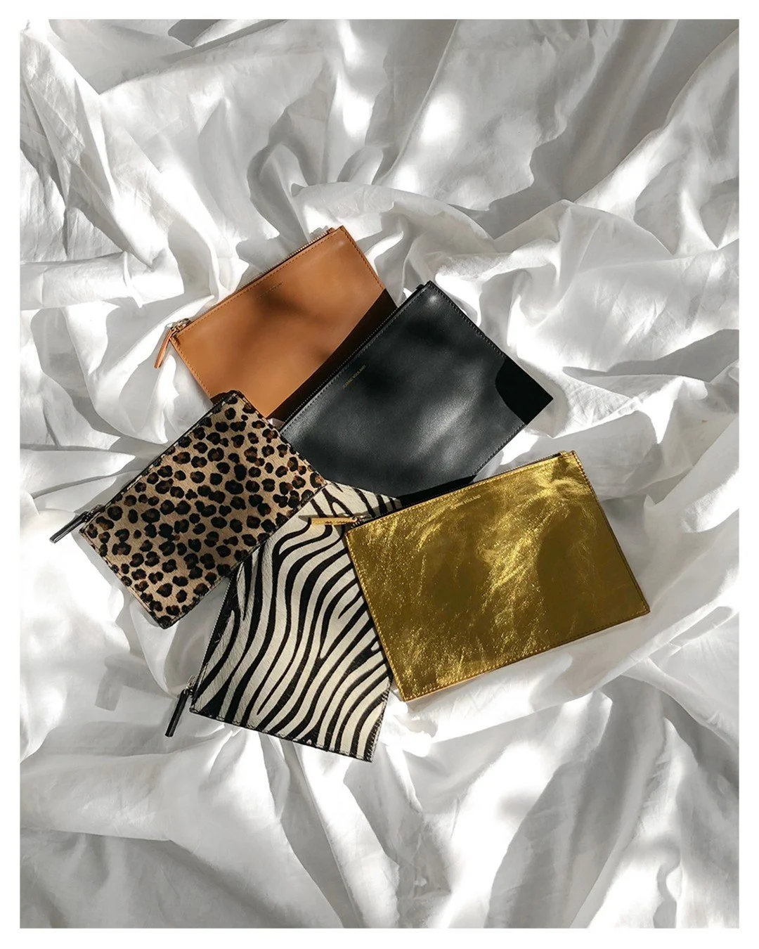 Cinq pochettes de différentes tailles et designs posées sur un drap blanc. Cataracte en cuir brun, pochette à motifs léopard, pochette à motifs zébré noir et blanc, pochette en cuir noir, et pochette en tissu métallisé doré.