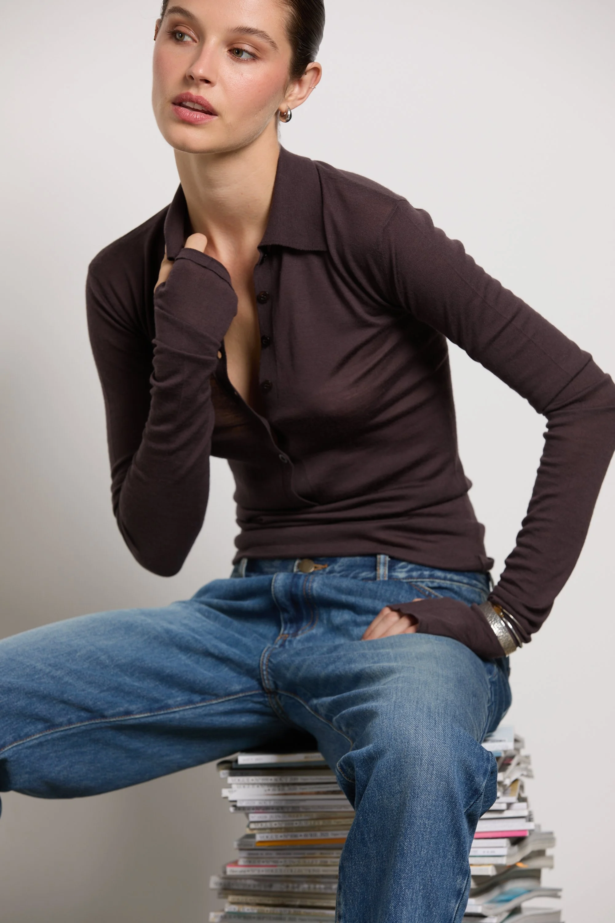 Une femme assise assise sur un tas de magazines blancs, portant un chemisier marron foncé et un jean bleu, regardant légèrement de côté.