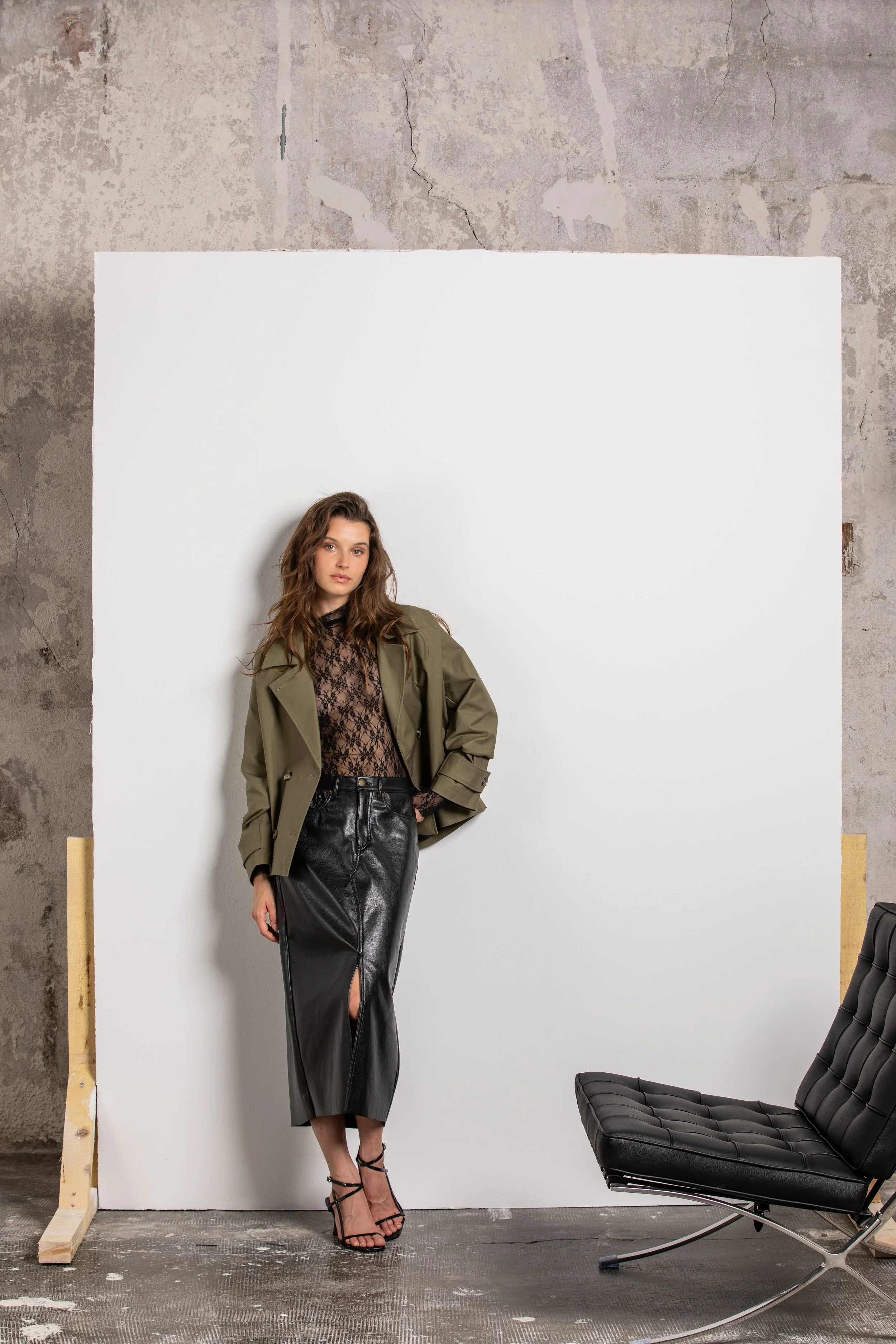 Une femme posant devant un fond blanc dans un studio avec un mur en pierre en arrière-plan, portant un trench kaki, une robe en dentelle noire, une jupe en cuir noir et des talons noirs à lanières.