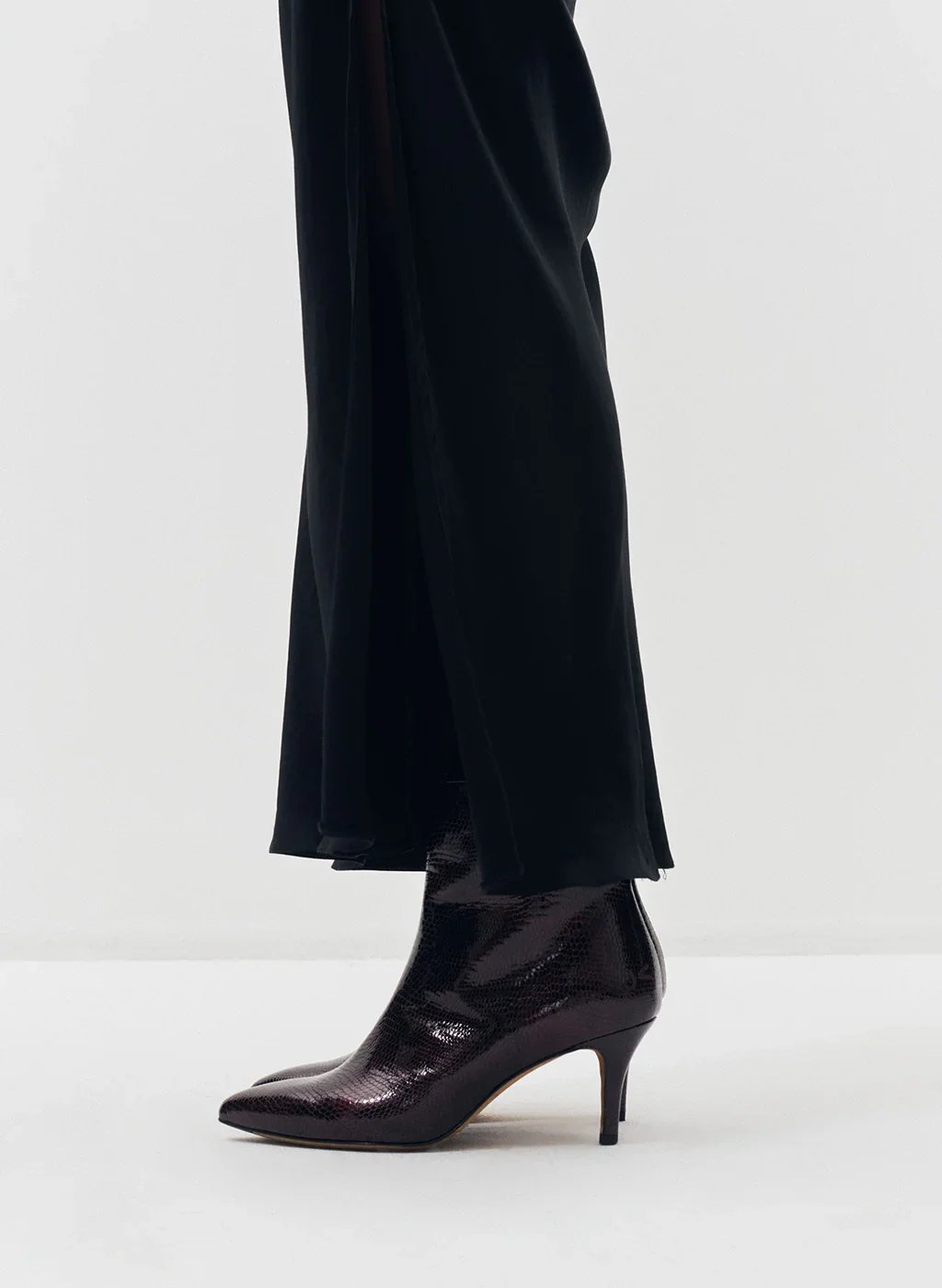 Une chaussure à talon élégante en cuir bordeaux avec une texture reptile, portée par une personne vêtue d'une longue jupe noire en satin.