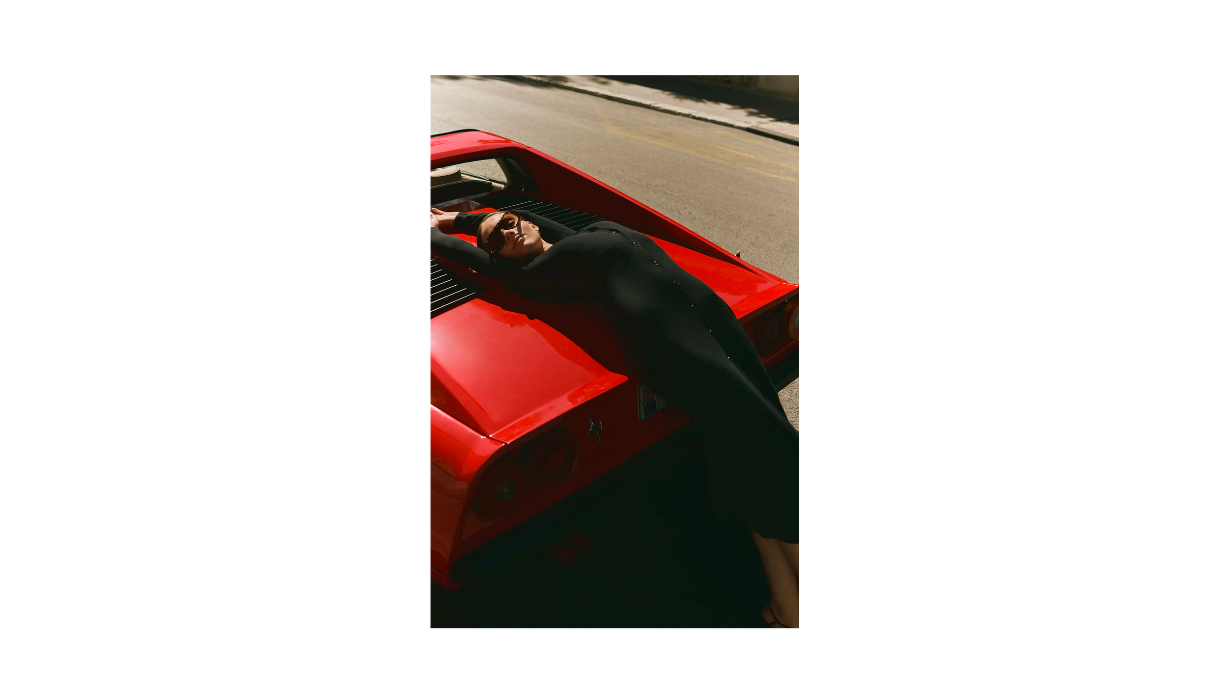 Une femme en noir allongée sur une voiture rouge, portant des lunettes de soleil, sur le trottoir.