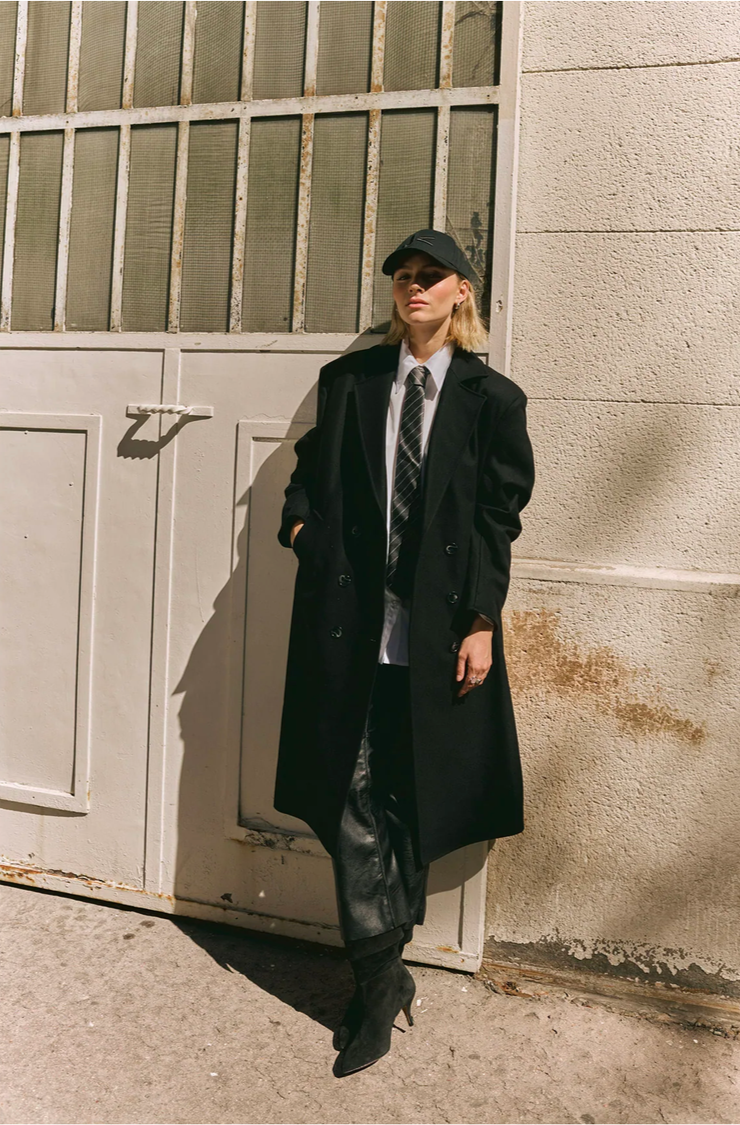 Jeune femme avec un manteau long noir, une chemise blanche, une cravate et un pantalon en cuir noir, portant une casquette noire, debout contre un mur en béton, dans un environnement urbain