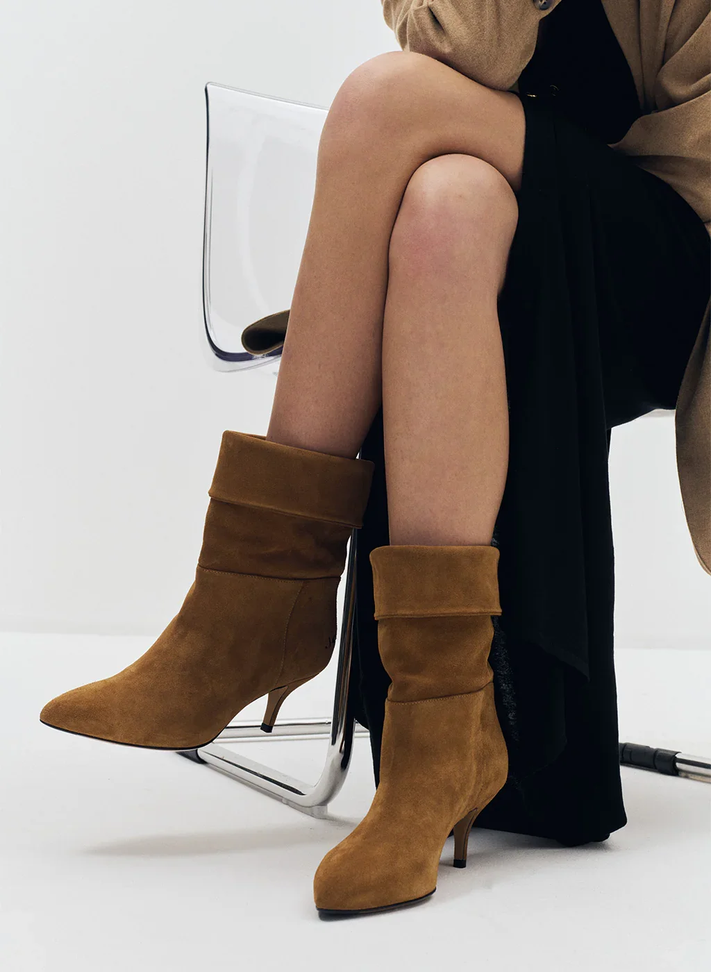 Une personne assise sur une chaise en verre, portant des bottes en suede marron et une jupe noire. La scène est minimaliste avec un fond blanc.