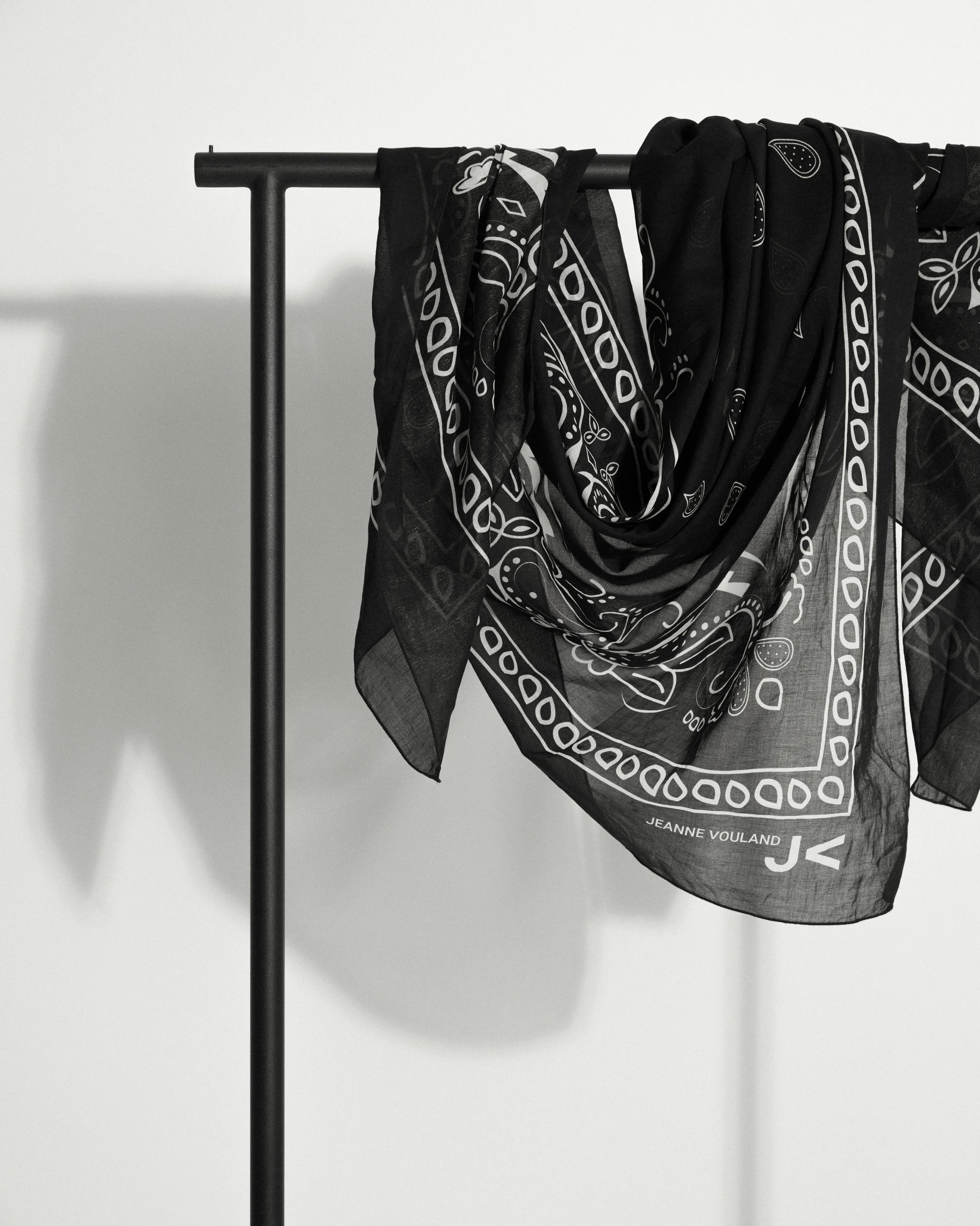 Un foulard noir avec un motif blanc accroché à un support en métal, avec l'inscription "Jeanne Voulard" et un logo en bas à droite.