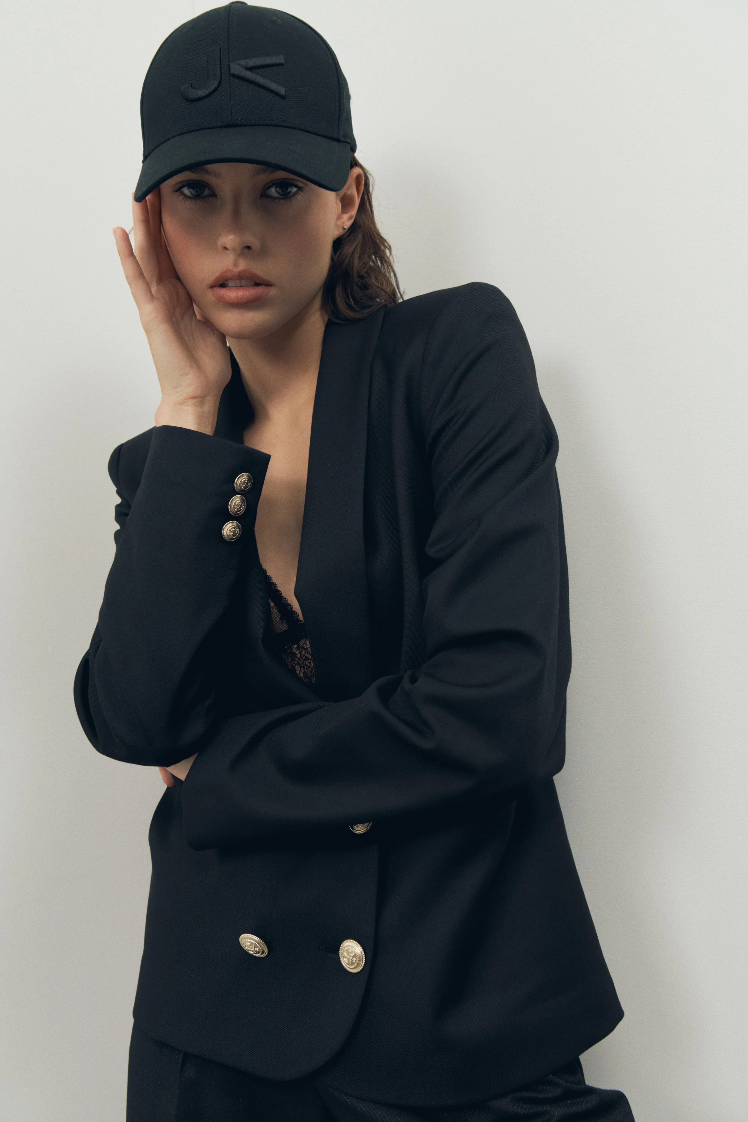 Jeune femme portant une casquette noire et un blazer noir avec boutons métalliques, posant contre un mur blanc.