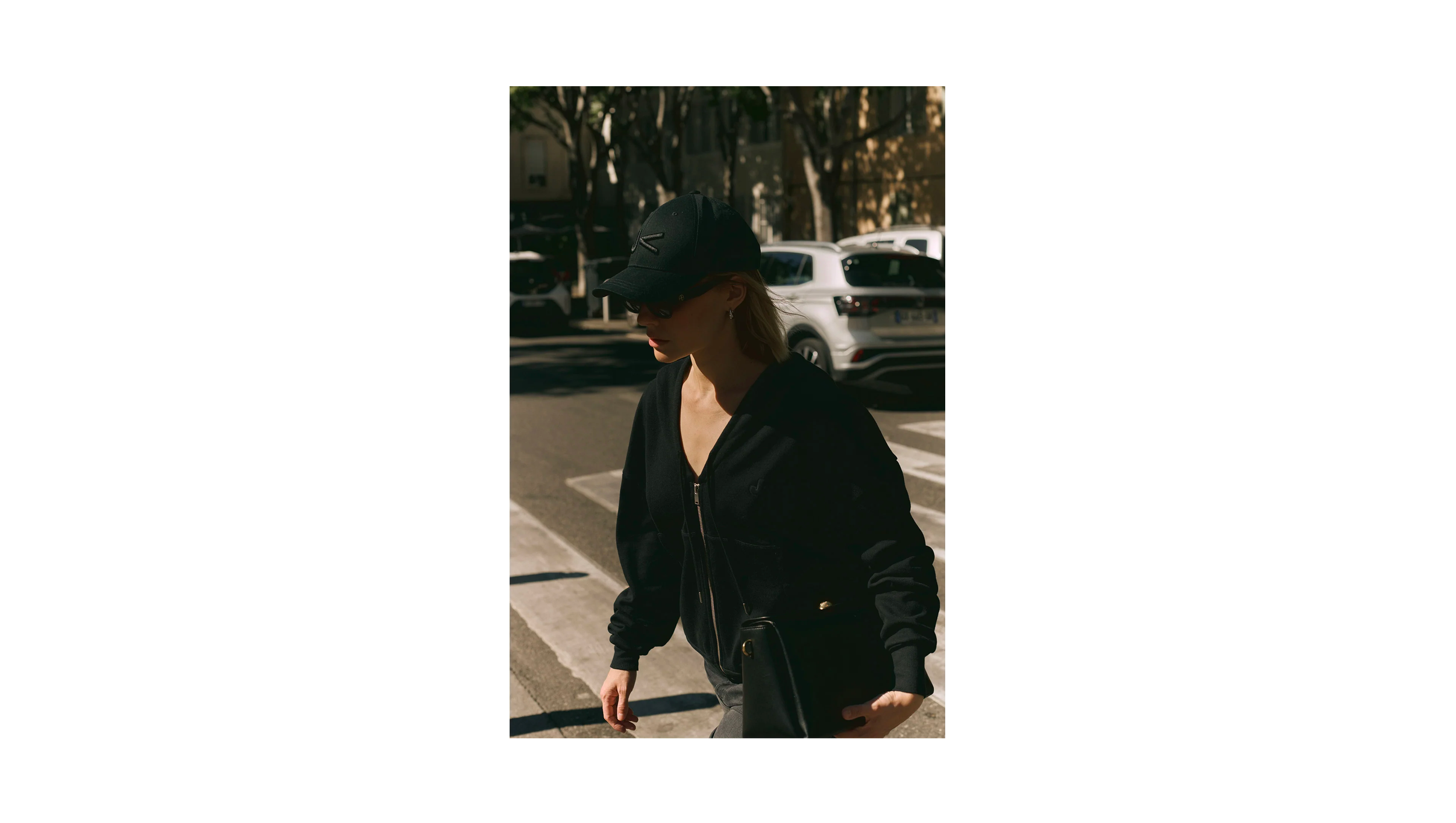 Une femme portant une casquette noire, des lunettes de soleil, un collier et un sac noir marche sur un passage pour piétons dans une ville pendant la journée. En arrière-plan, des voitures sont stationnées et des arbres ombragent la scène.