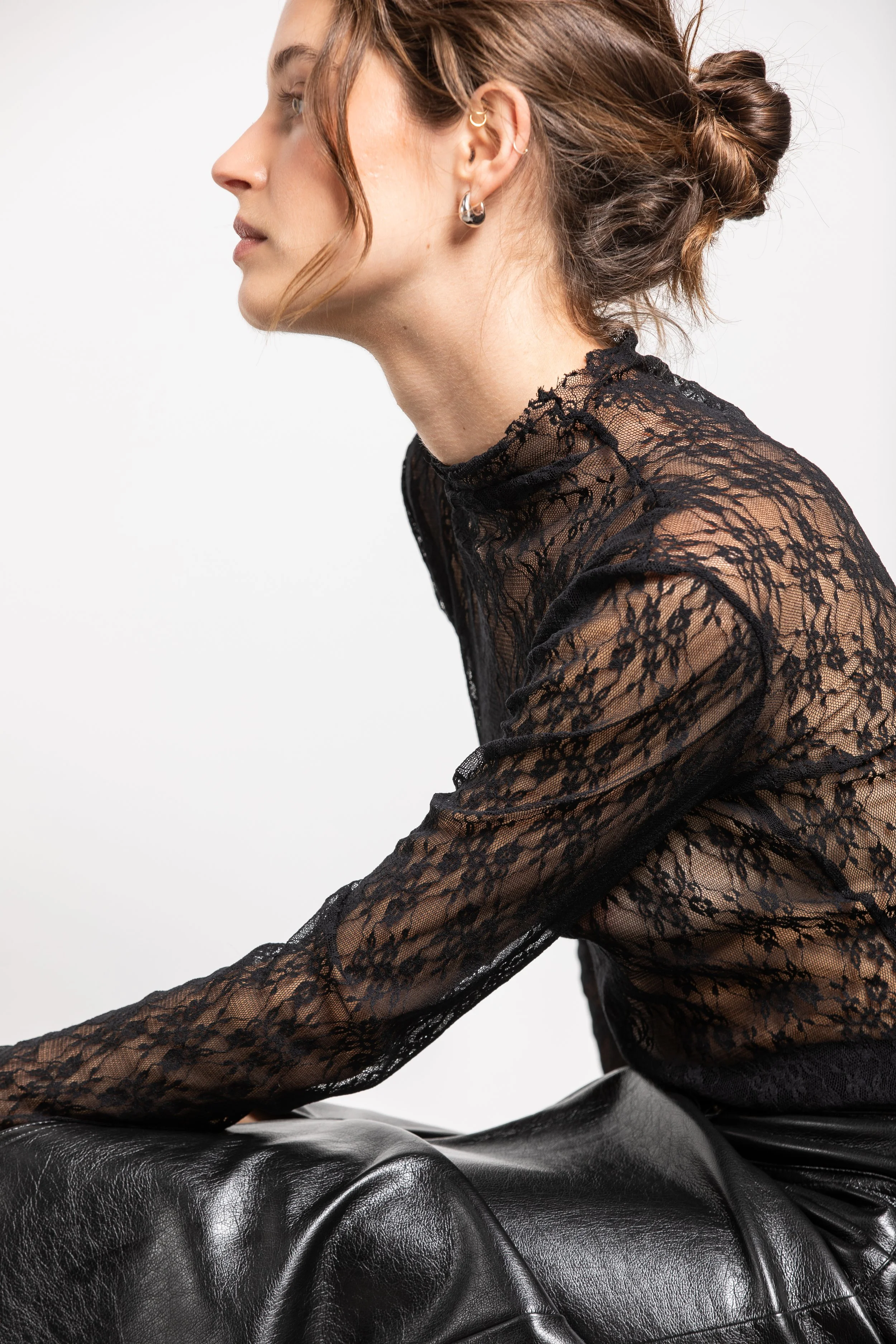 Profil d'une femme assise portant un haut en dentelle noir et un pantalon en cuir noir, avec des boucles d'oreilles en argent.