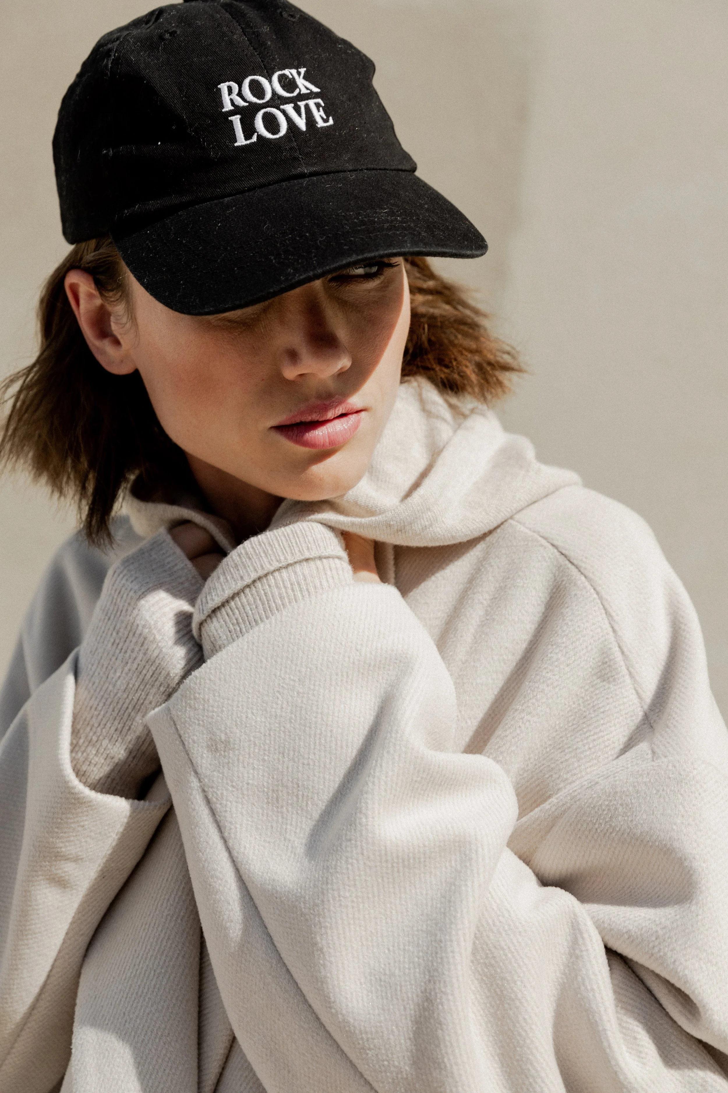 Une femme portant une casquette noire avec l'inscription "ROCK LOVE" en blanc, un sweat à capuche beige et une paire de gants gris clair, regardant vers le bas avec une expression pensive.