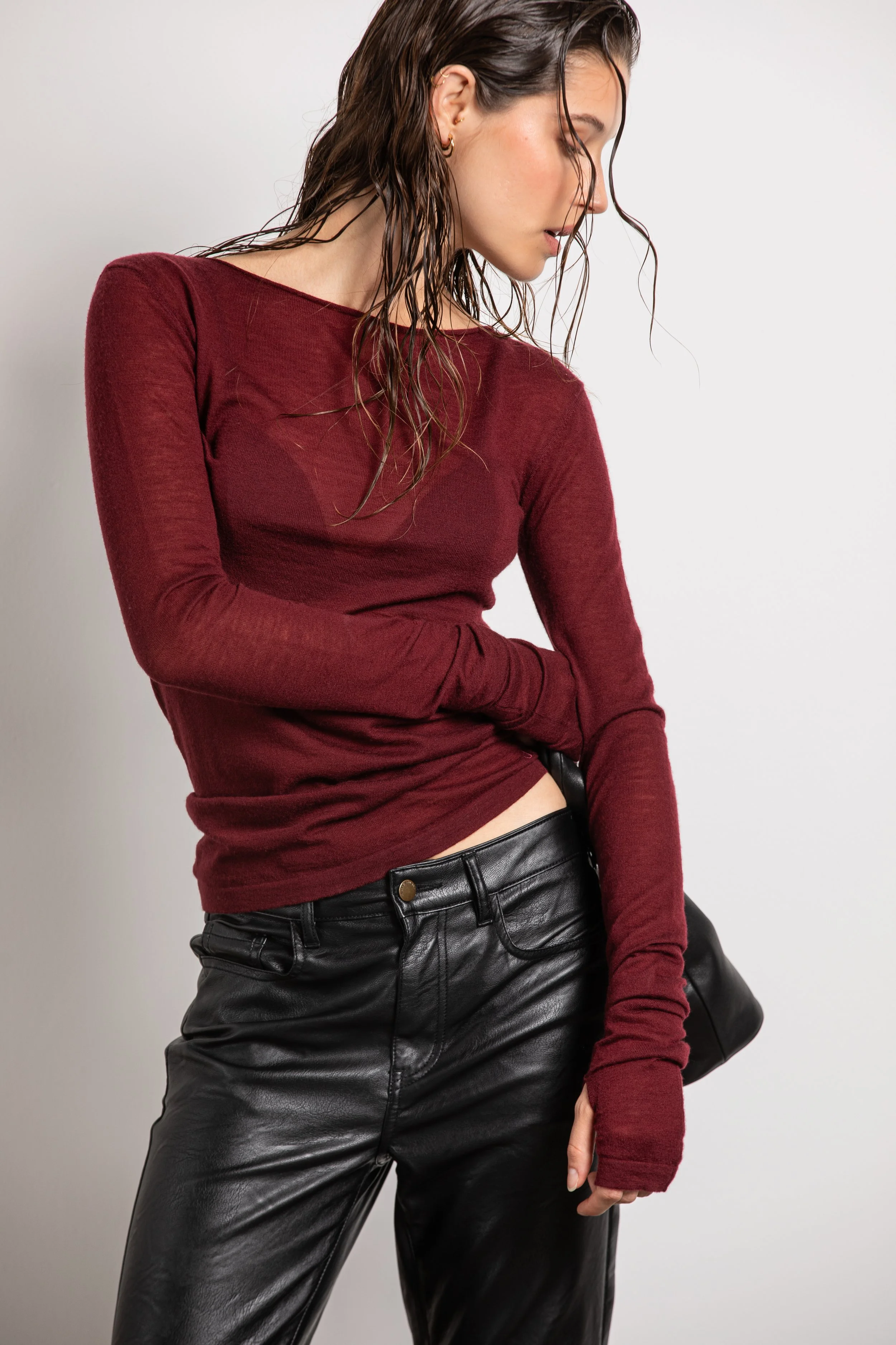 Une femme portant un haut rouge foncé en manches longues et un pantalon en cuir noir, avec des cheveux bouclés légèrement mouillés, se tenant contre un fond blanc