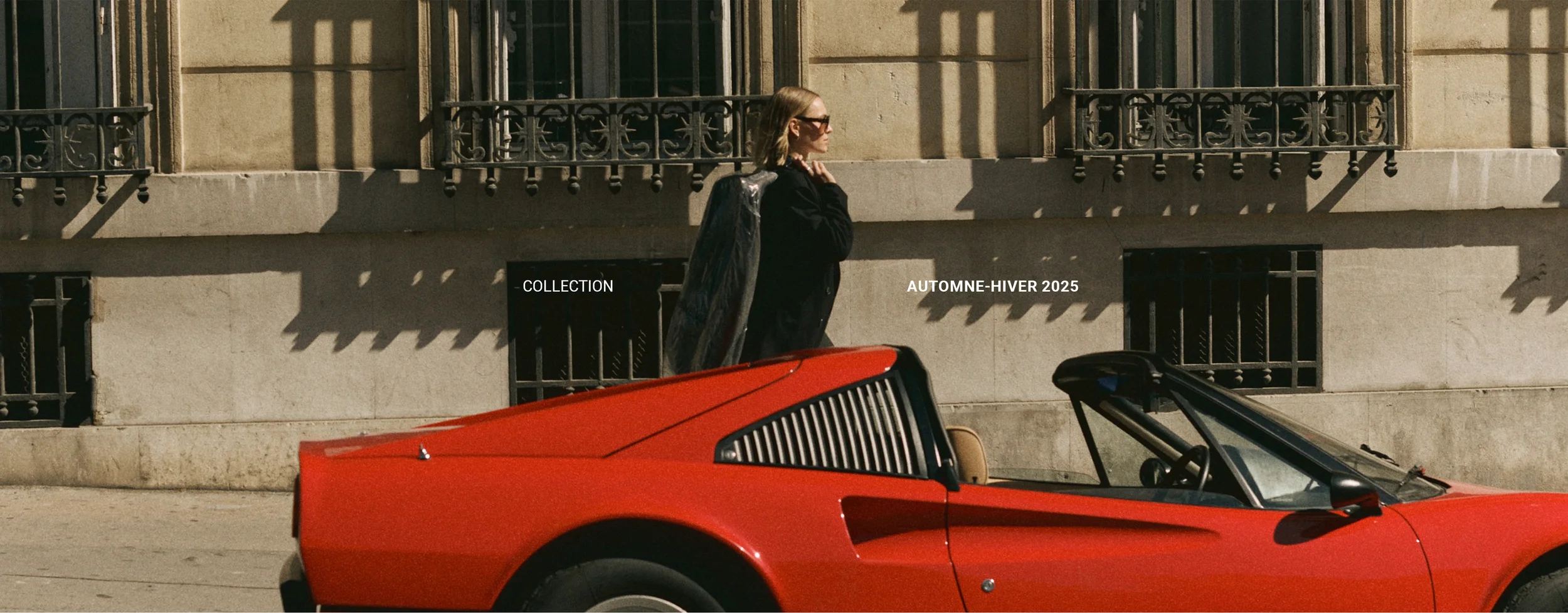 Une femme portant des lunettes de soleil et un manteau noir marche à côté d'une voiture rouge décapotable stationnée devant un bâtiment en pierre.