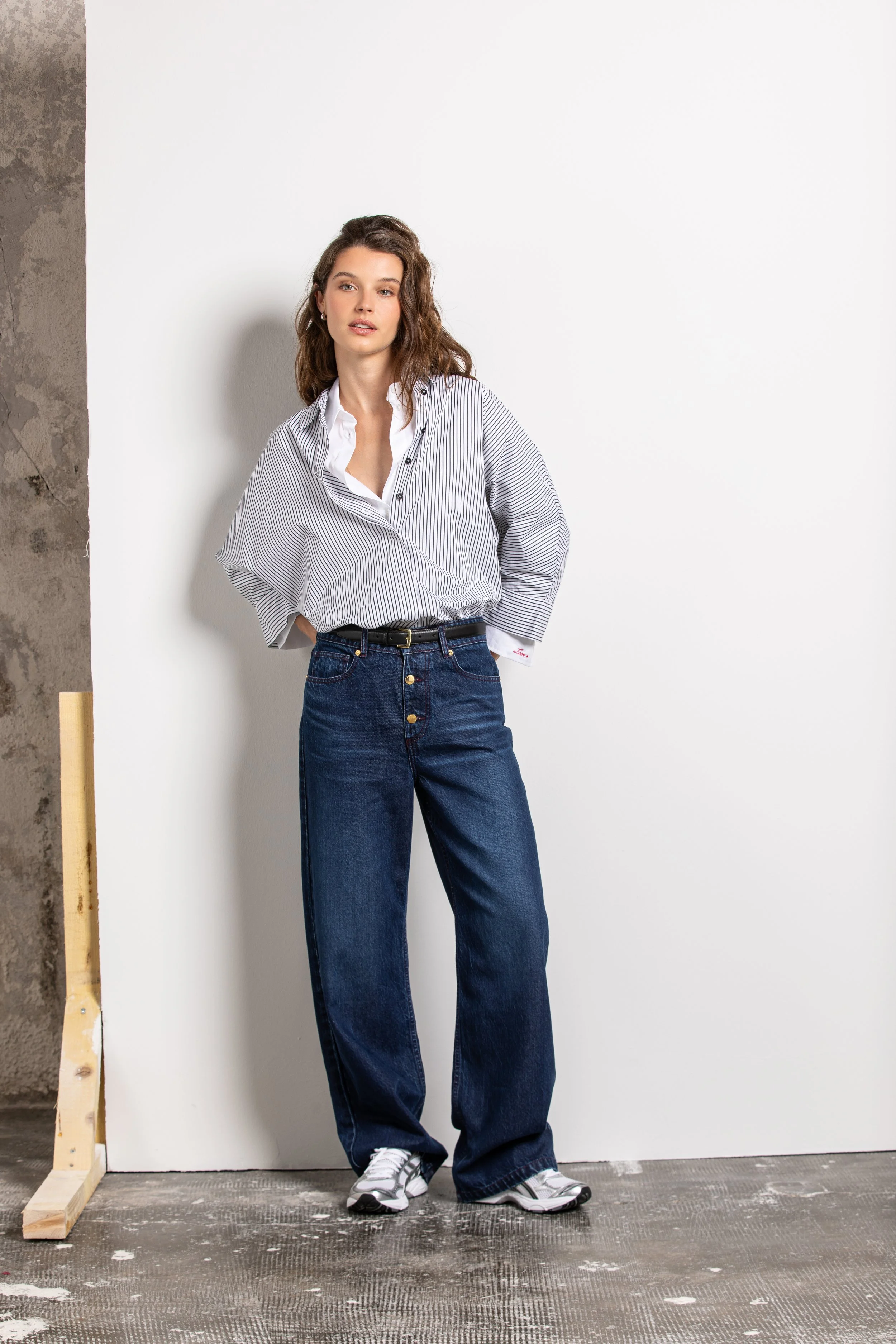 Jeune femme avec chemise à rayures Bleu et blanc, jean ample et chaussures de sport, posant contre un mur blanc