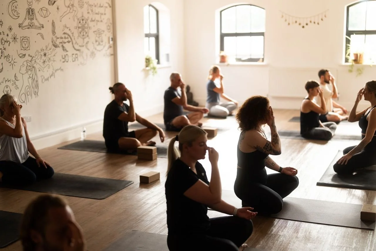 Minimalist interior with a wooden chair, a floating wooden shelf with a book and a small object, and a wooden trash bin against a plain textured wall and wooden flooring.