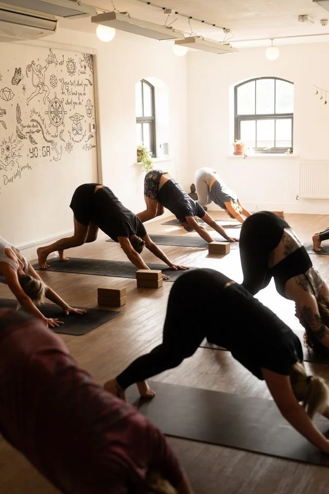 Minimalist room with wooden furniture, including a desk, chair, and floating shelf on a beige wall, with a small wooden waste bin on the floor.