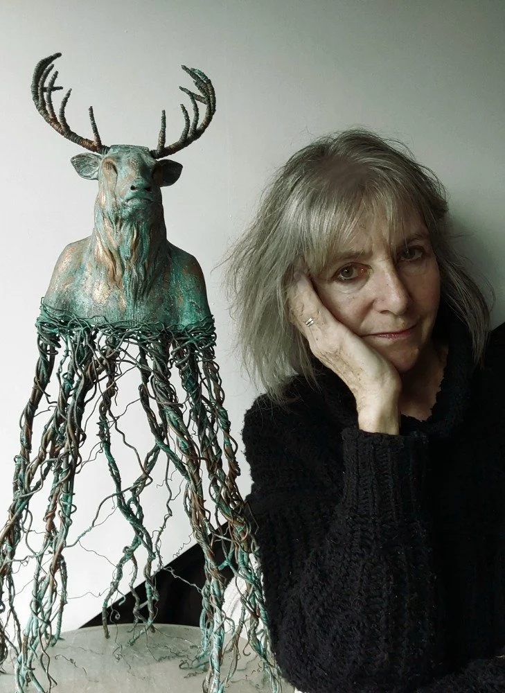 A woman with shoulder-length gray hair, wearing a black sweater, resting her head on her hand, is sitting next to a sculpture of a deer with antlers. The sculpture's body is creatively made of twisted, wire-like roots or branches.