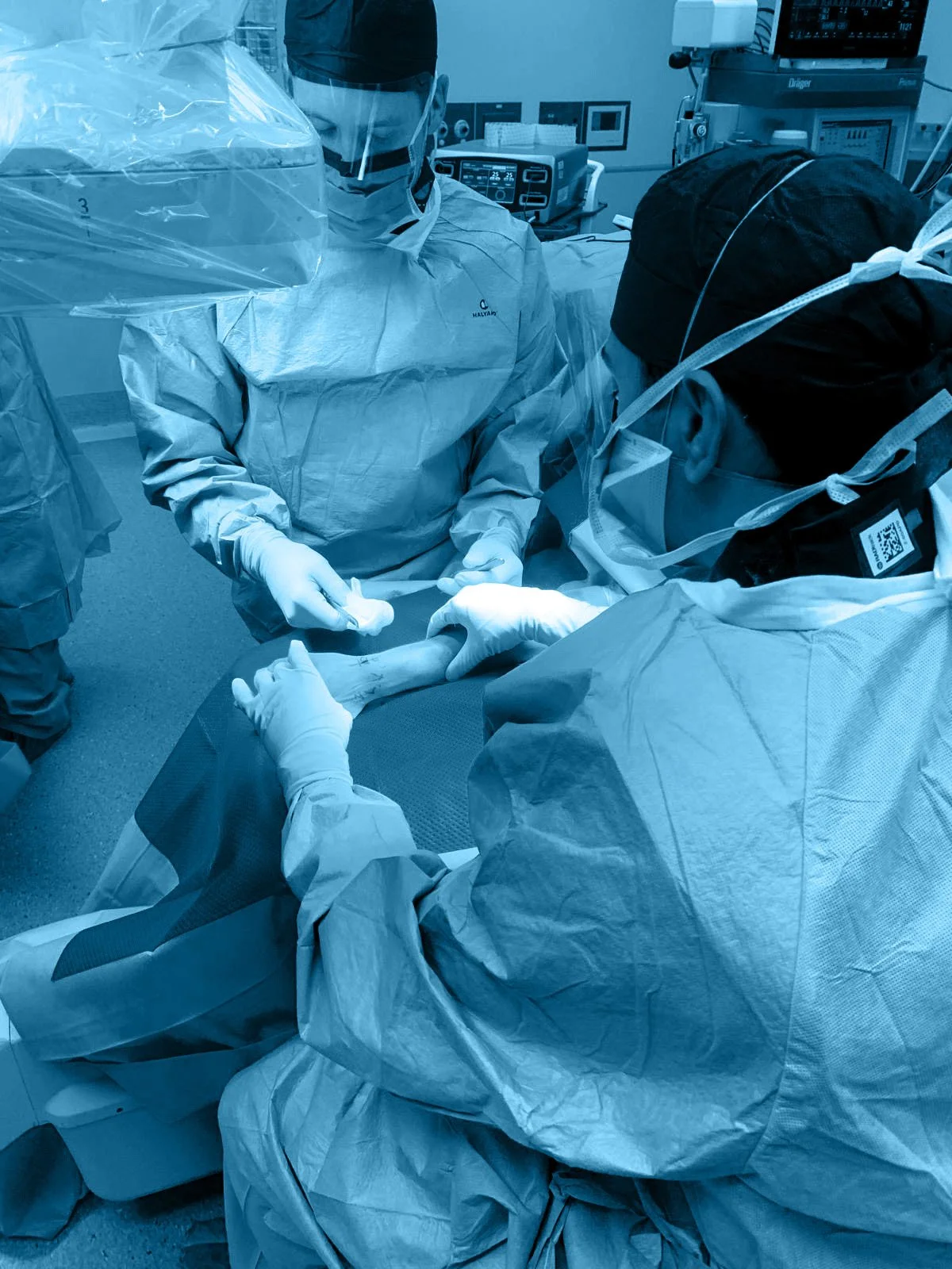 Medical professionals in surgical gowns, masks, and gloves perform a procedure on a patient's arm in an operating room with medical equipment and monitors in the background.