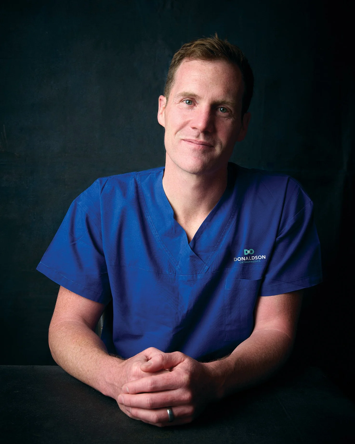 Matthew Donaldson, Orthopaedic Surgeon, wearing blue medical scrubs sitting with his hands folded, smiling at the camera against a dark background.