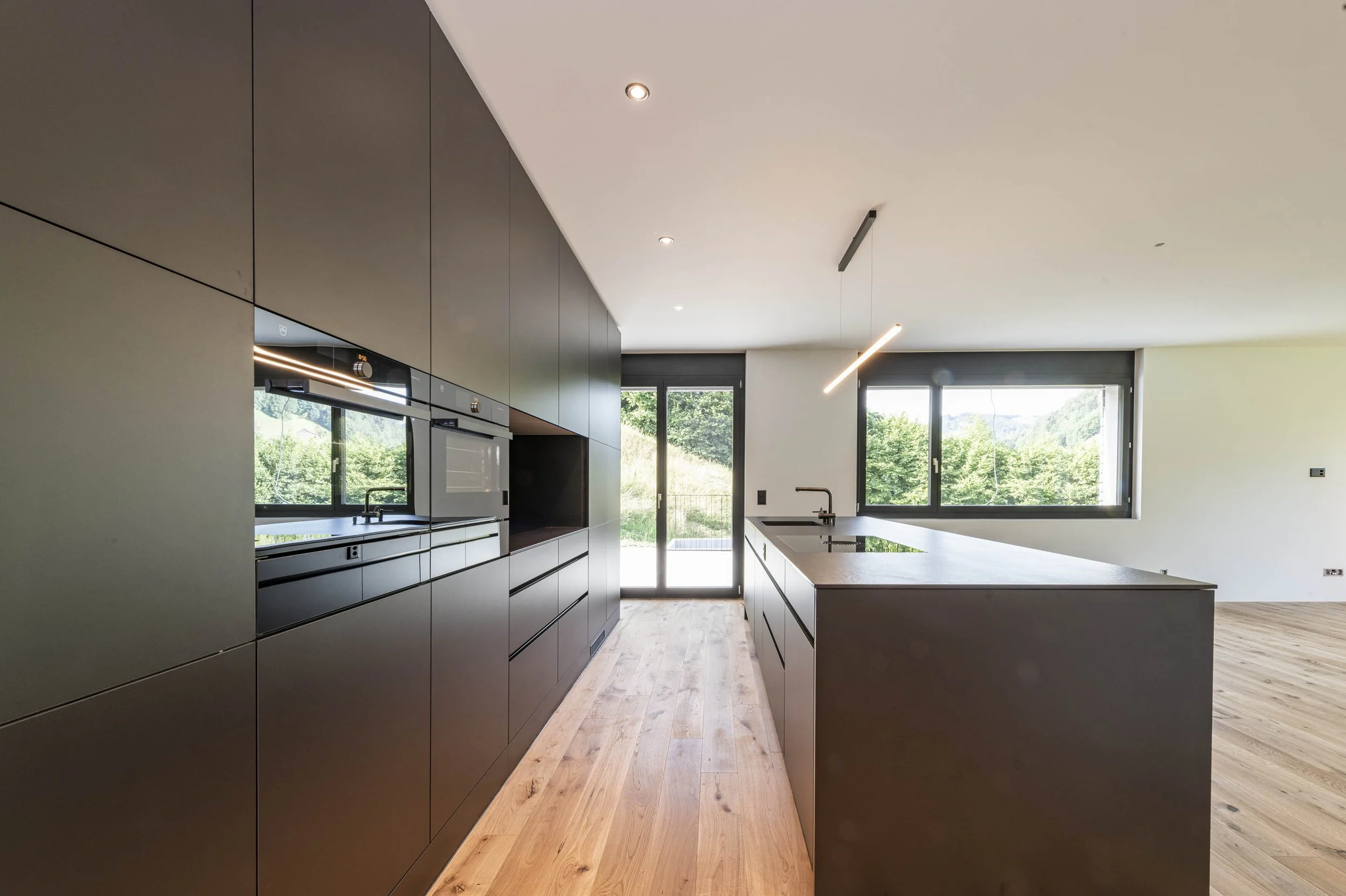 Moderne, minimalistische Küche mit schwarzen Schränken, einer Kücheninsel, großen Fenstern und Holzboden.