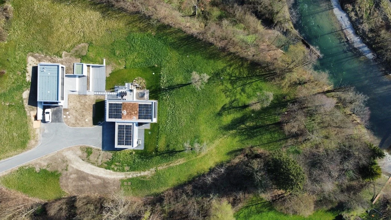 Vogelperspektive auf ein modern gebautes Haus mit Solaranlagen auf dem Dach, umgeben von grünem Garten, Bäumen und einem Fluss im Hintergrund.