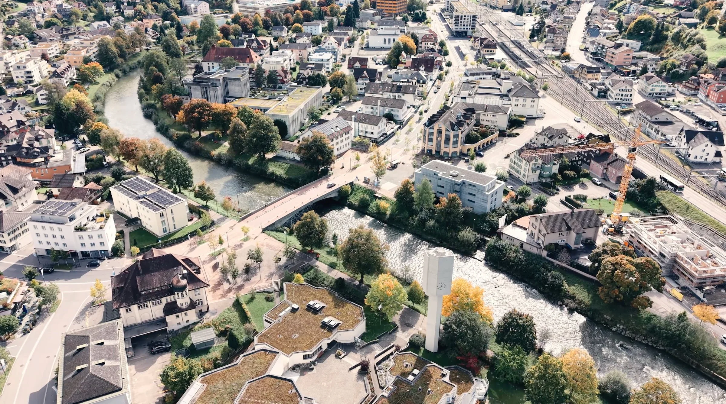 Luftaufnahme einer Stadt mit Fluss, Brücken, Wohnhäuser, Baustellen und Straßen, umgeben von Bäumen.